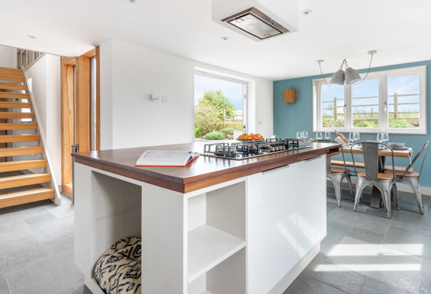 Modern kitchen and dining area with large window and sliding door, wooden staircase, white and teal walls, island with cookbook, and dining table set with glasses and plates.