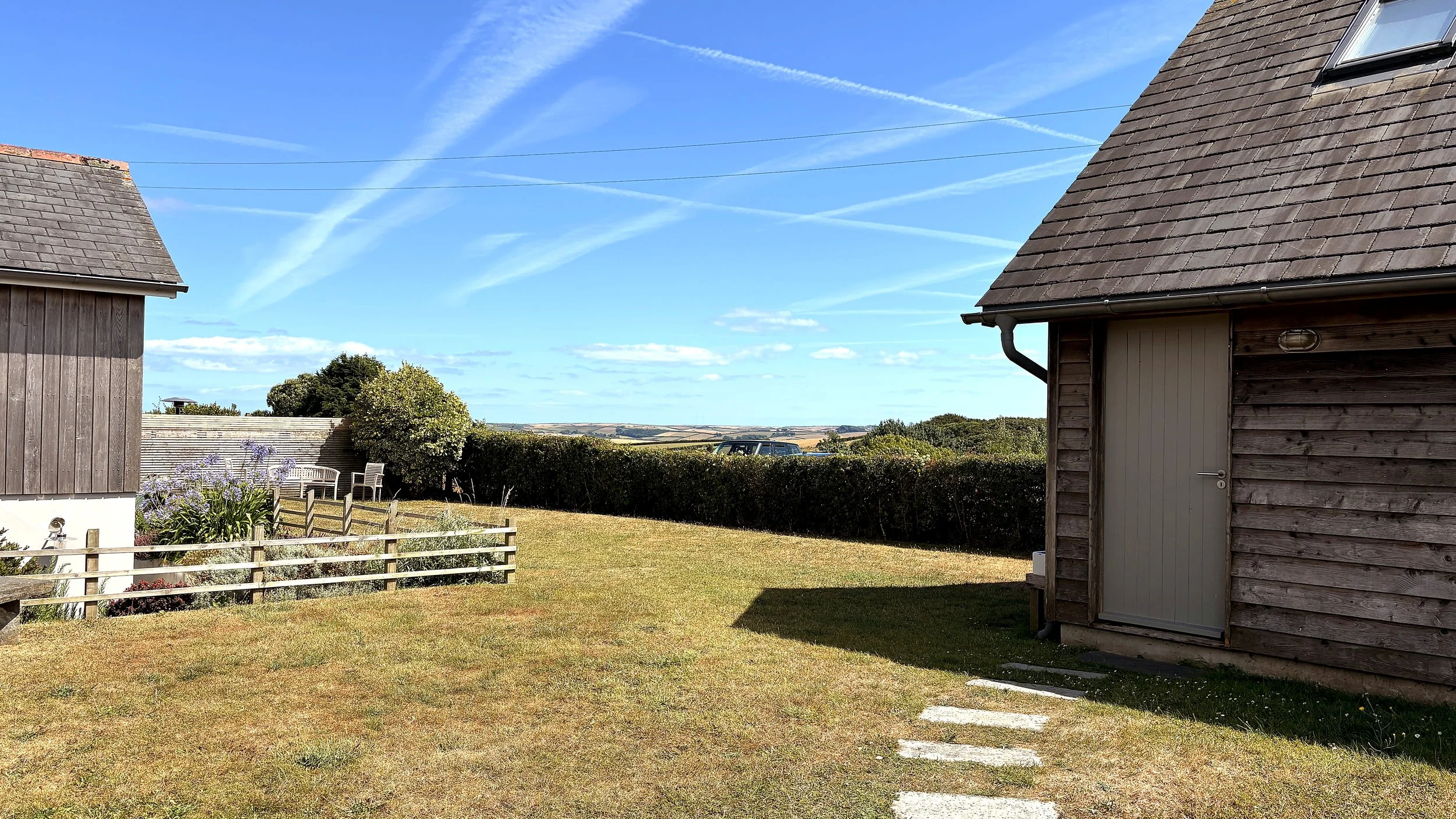 A backyard scene with two wooden buildings, a fence with flowers, and a view of distant fields under a blue sky with contrails.