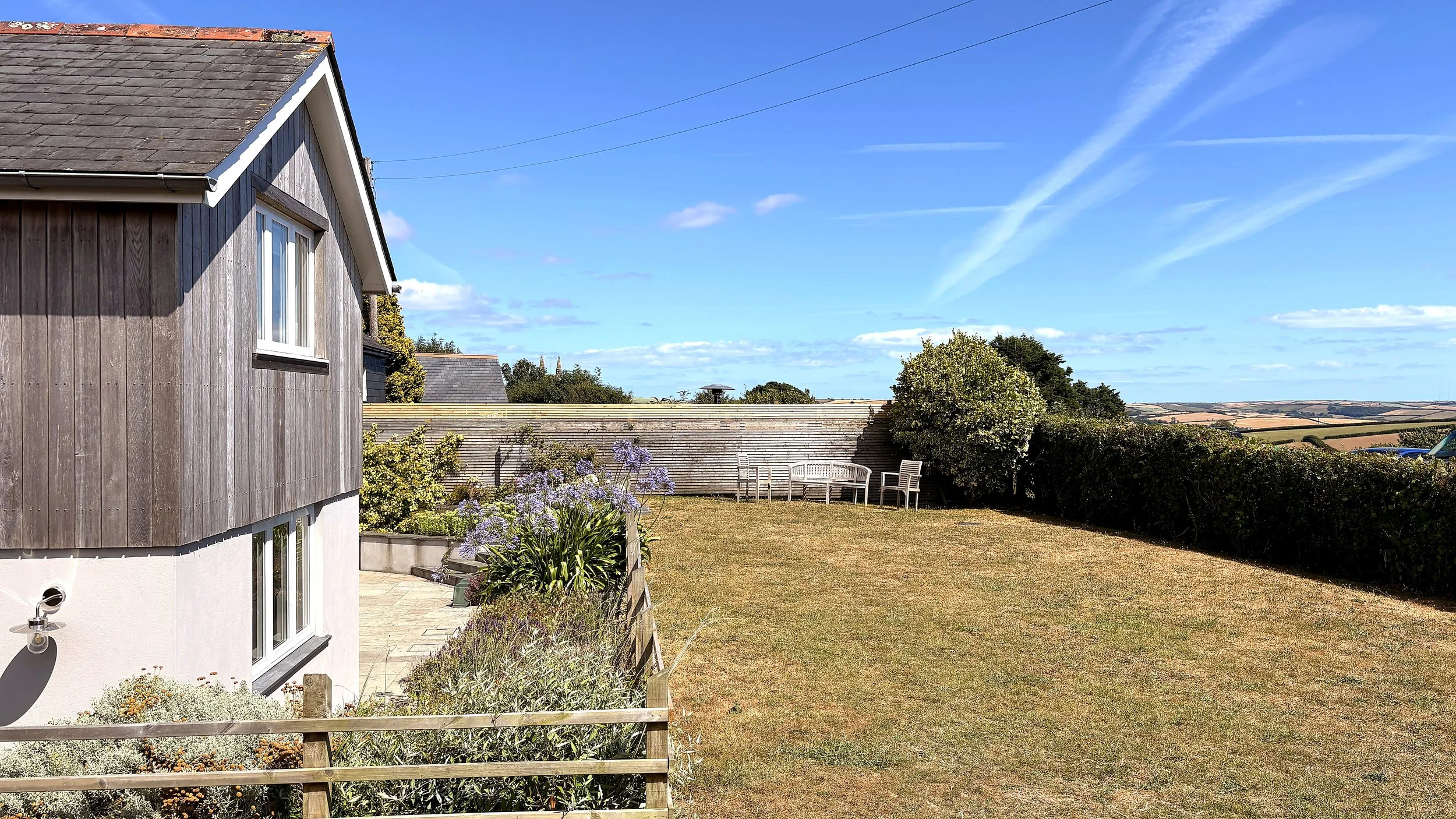 A backyard with a lawn, a garden bed with flowers, a patio, a neighboring house, and a view of rolling hills under a blue sky with some clouds.