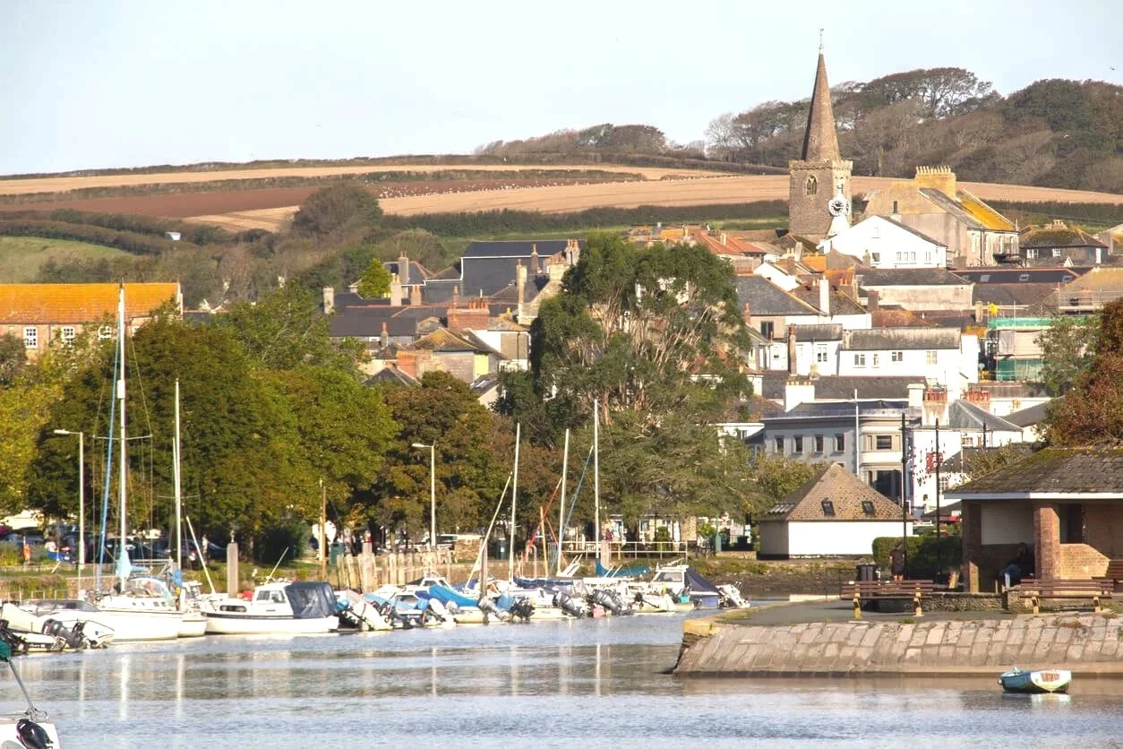 A harbor with boats docked, green trees, and buildings behind, with a church steeple in a small town set against rolling hills in the background.