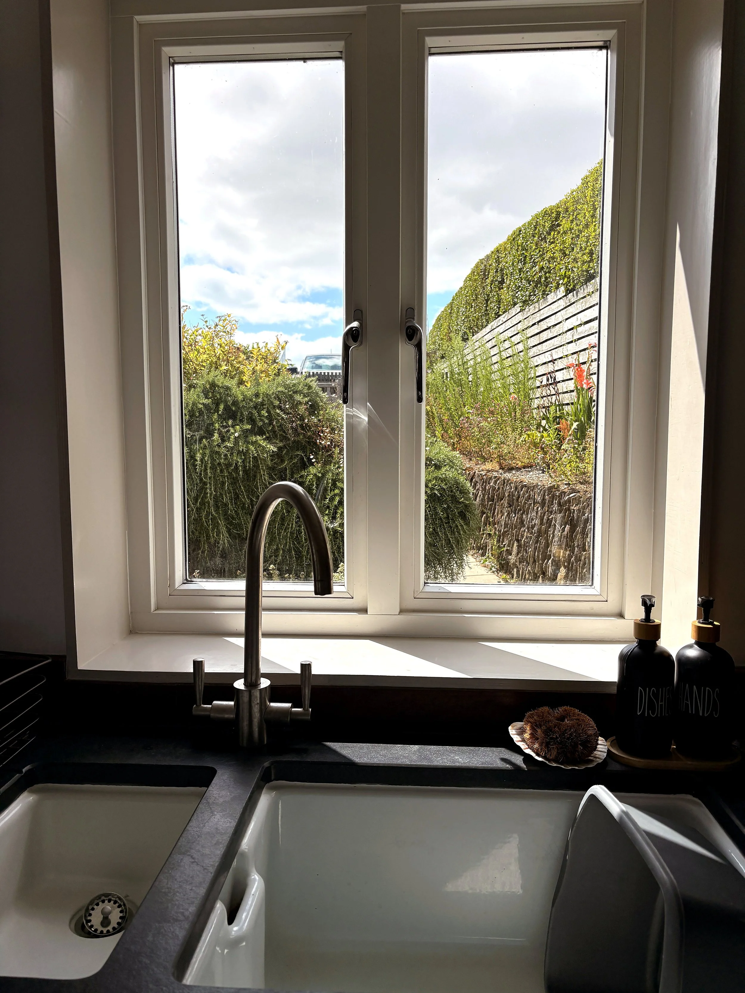 A kitchen with a window view of a garden, two white sinks, a black countertop, and a dish soap dispenser on the right side.