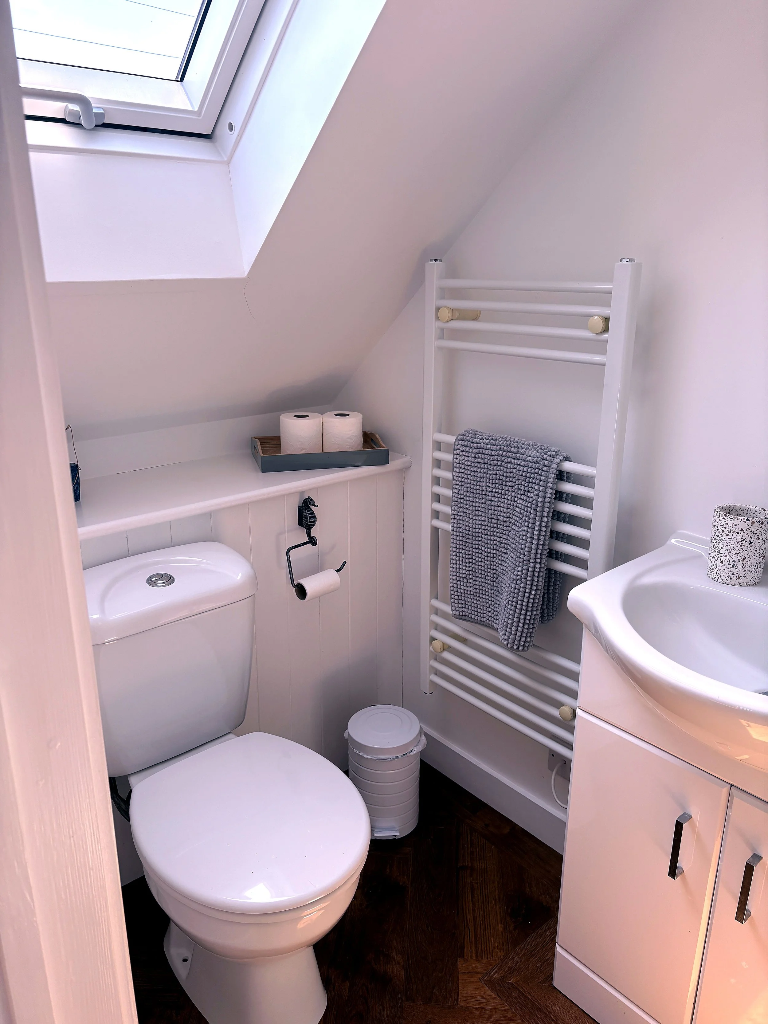 Small bathroom with a toilet, a white cabinet, a small sink, a window on the slanted ceiling, a towel radiator with a gray towel, three toilet paper rolls on a tray, and a small trash bin.