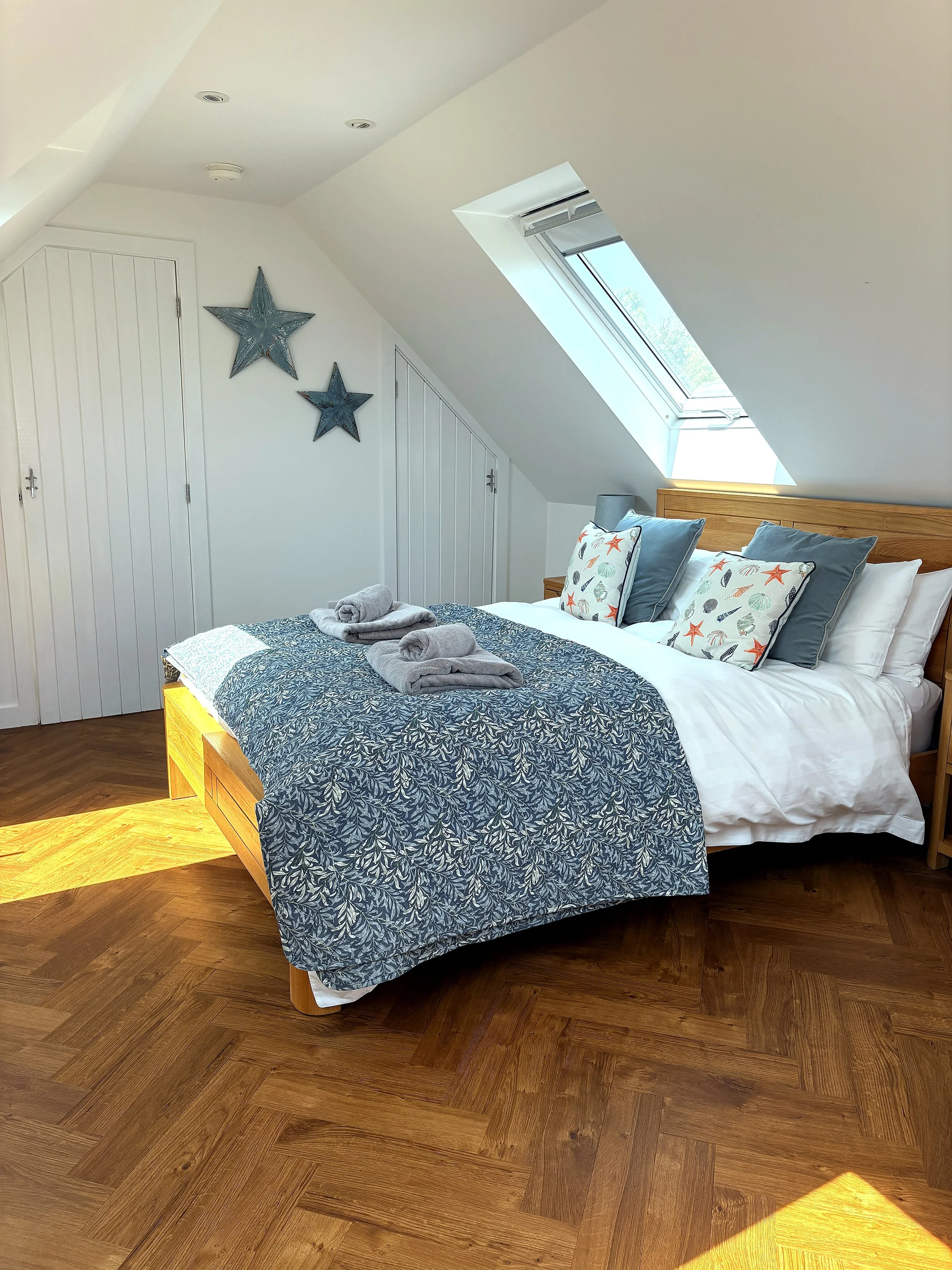A bedroom with a bed, decorative pillows with seashell and starfish designs, and towel arrangements on top. There are two dark blue star wall decorations and a skylight window on the sloped ceiling, allowing natural light into the room. The flooring 