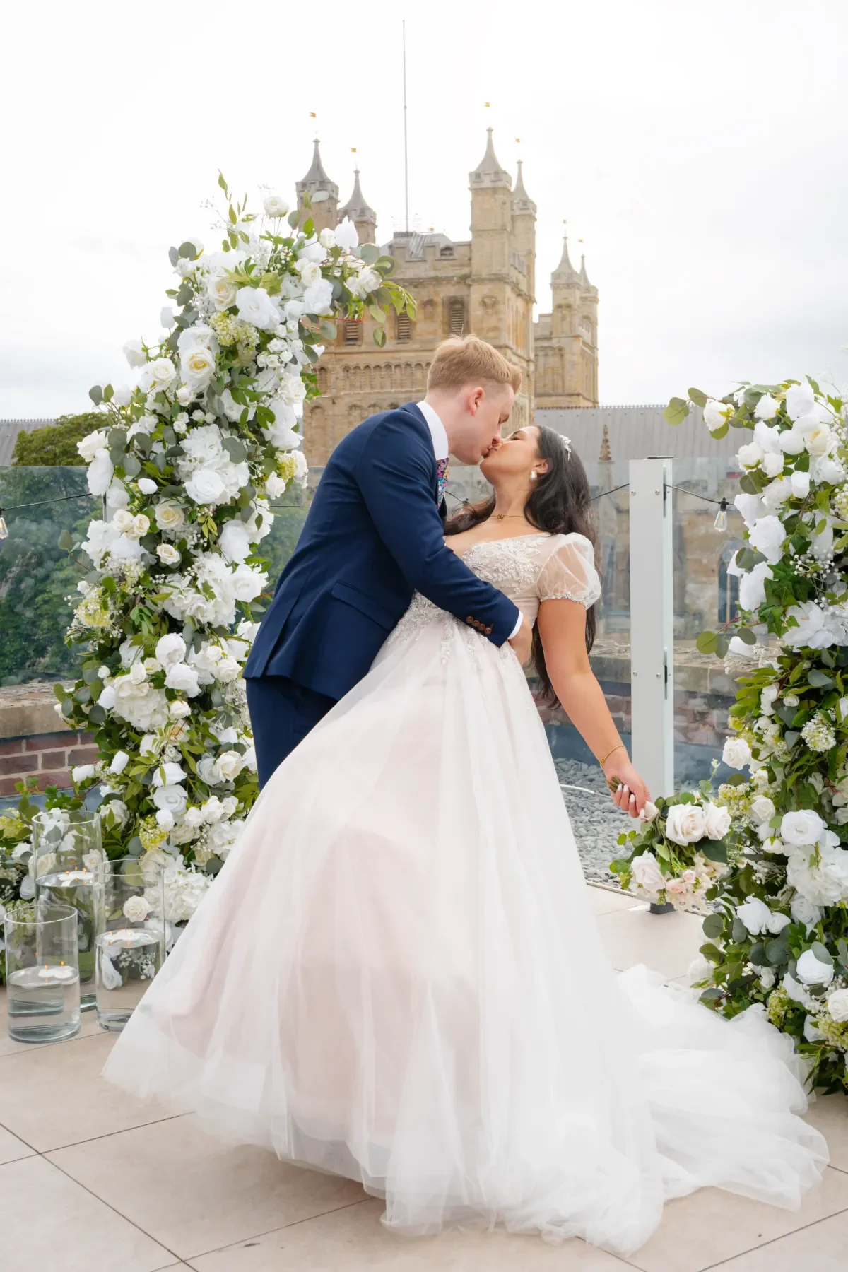 A bride and groom kissing during their wedding ceremony outdoors in front of a Exeter Cathedral, decorated with white floral arrangements.