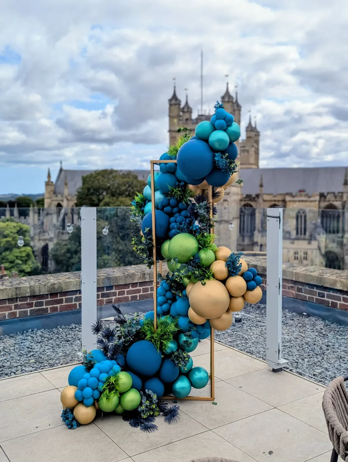 A decorative balloon arrangement with blue, green, beige, and black balloons, complemented by greenery and floral accents, set against a backdrop of a Exeter Cathedral on a cloudy day.