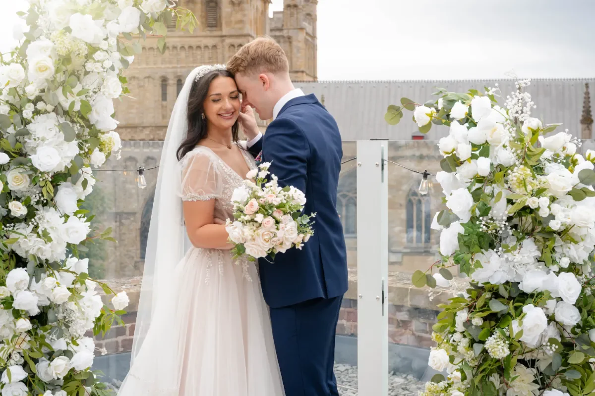 A bride and groom are kissing under an arch of white flowers during their wedding, with a historic building in the background.