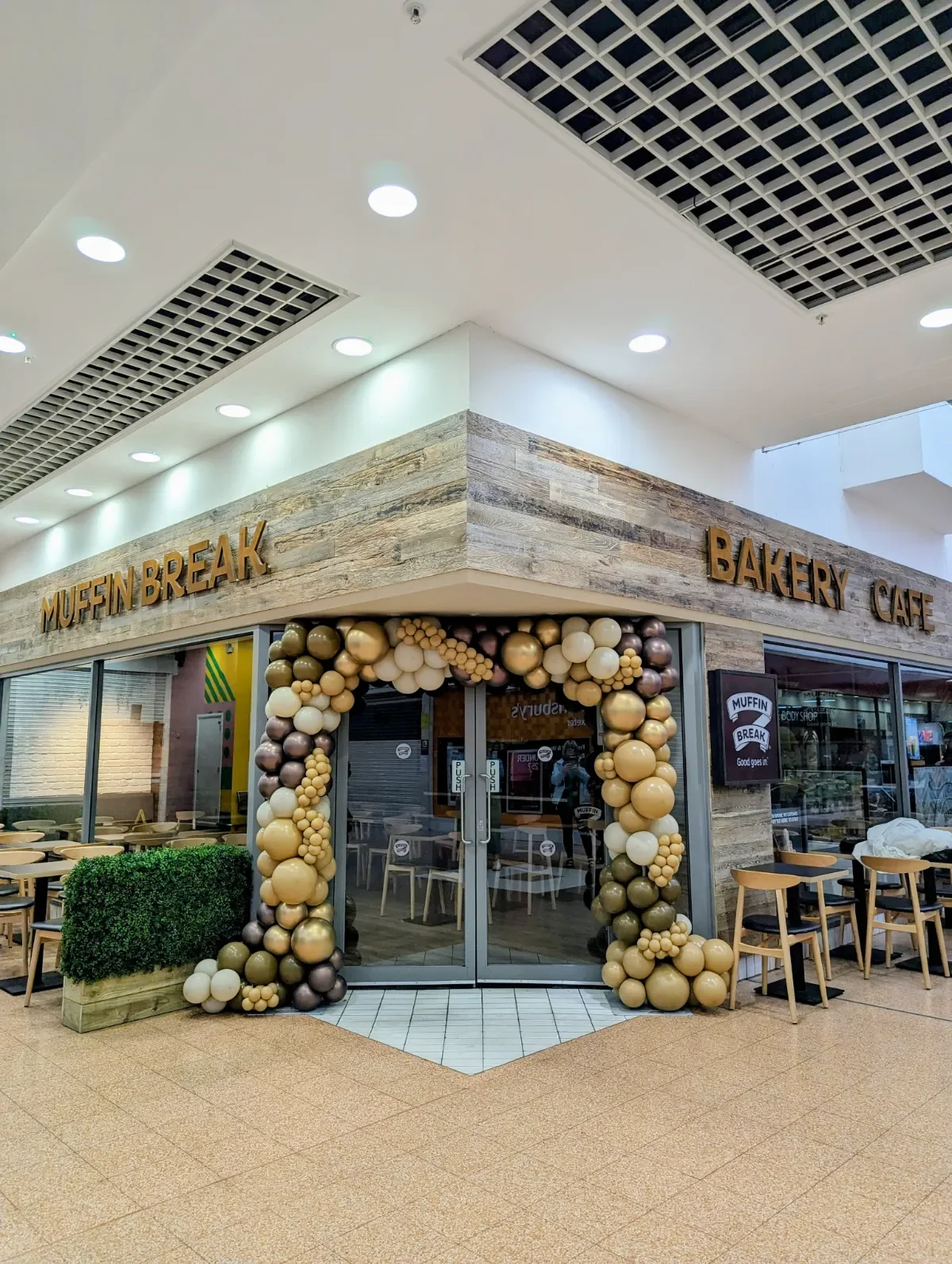 Restaurant entrance decorated with a balloon arch in beige, brown, and cream colors, featuring a sign with gold letters reading 'MUFFIN BREAK' and additional sign saying 'BAKERY CAFE' on the wooden exterior wall.