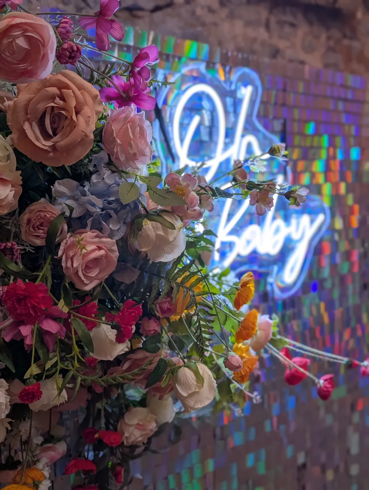 A colorful flower arrangement of roses and other flowers in front of a neon sign with the word "Baby" in blue and white, against a brick wall background.