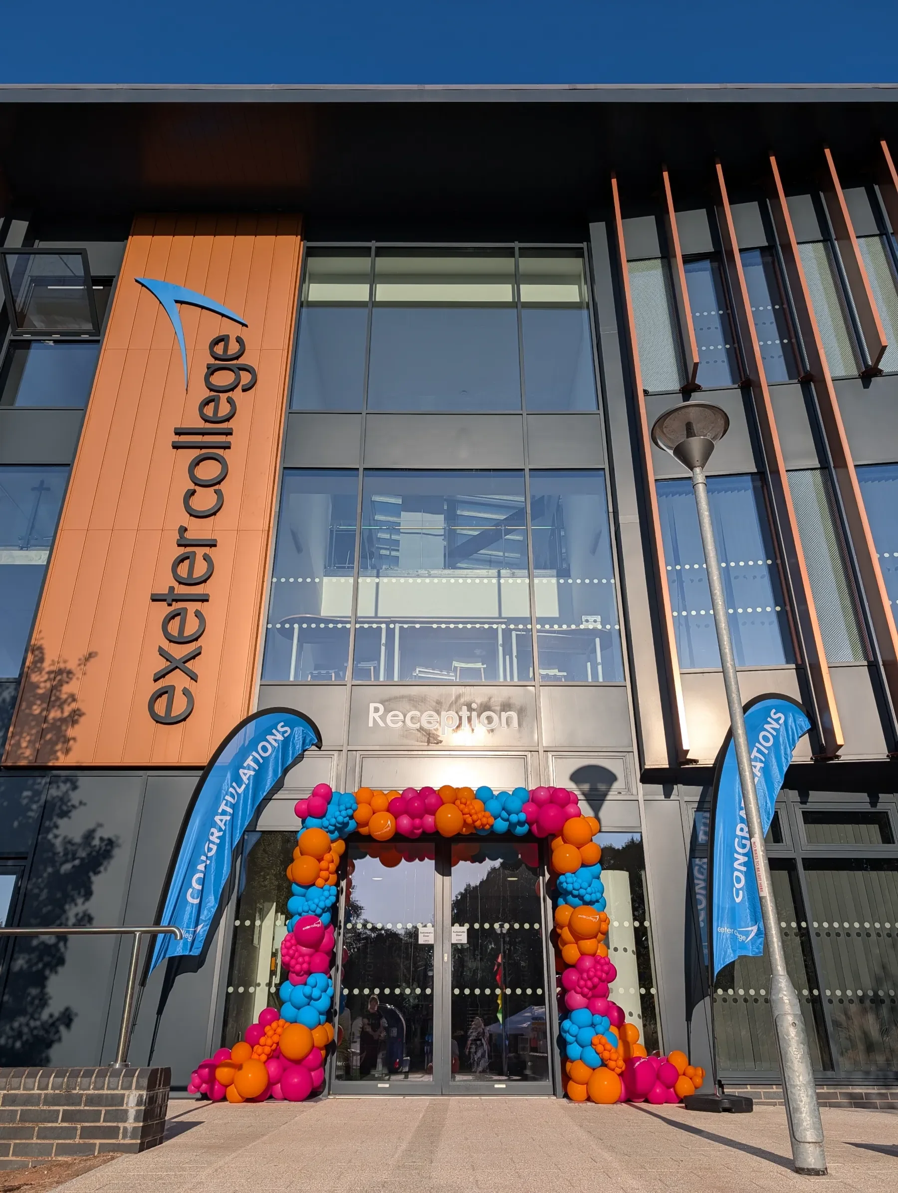 Modern building with glass windows and wooden accents. Balloons in pink, orange, and blue form an arch at the entrance. Two blue flags with white text stand on either side of the entrance. The building has signage indicating it is the reception area for an event or celebration.