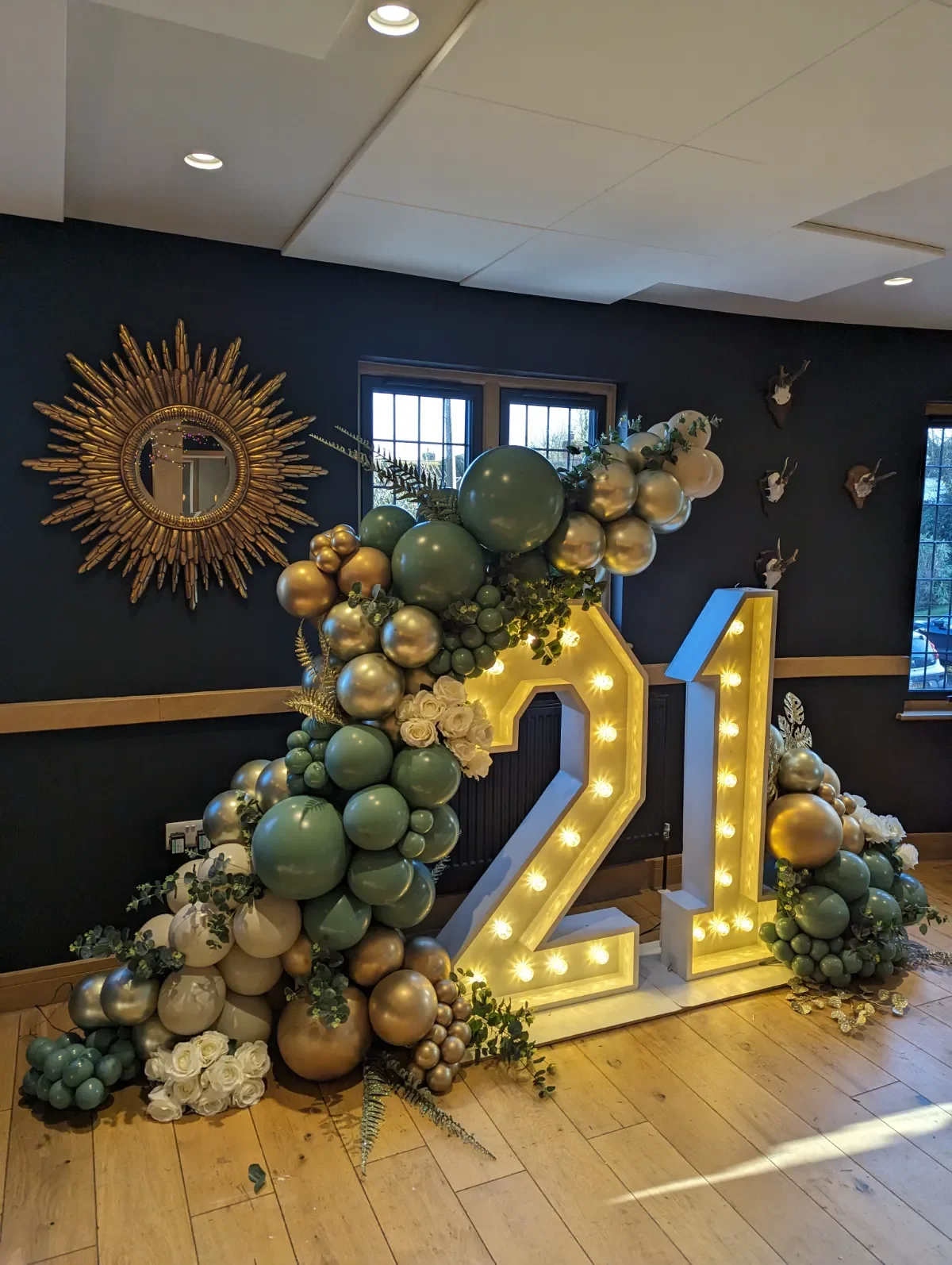 Decorative display with illuminated number "21" surrounded by balloons in green, gold, and white, with flowers and greenery, set against a dark wall with a gold sunburst mirror.