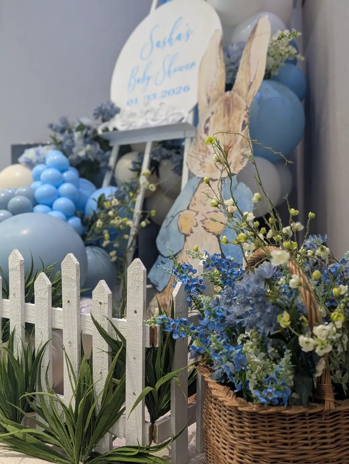 Decoration for a baby shower with blue and white balloons, flowers, a small white picket fence, a wicker basket with blue flowers, and a sign that says 'Sarah Baby Shower 01.09.2026'.