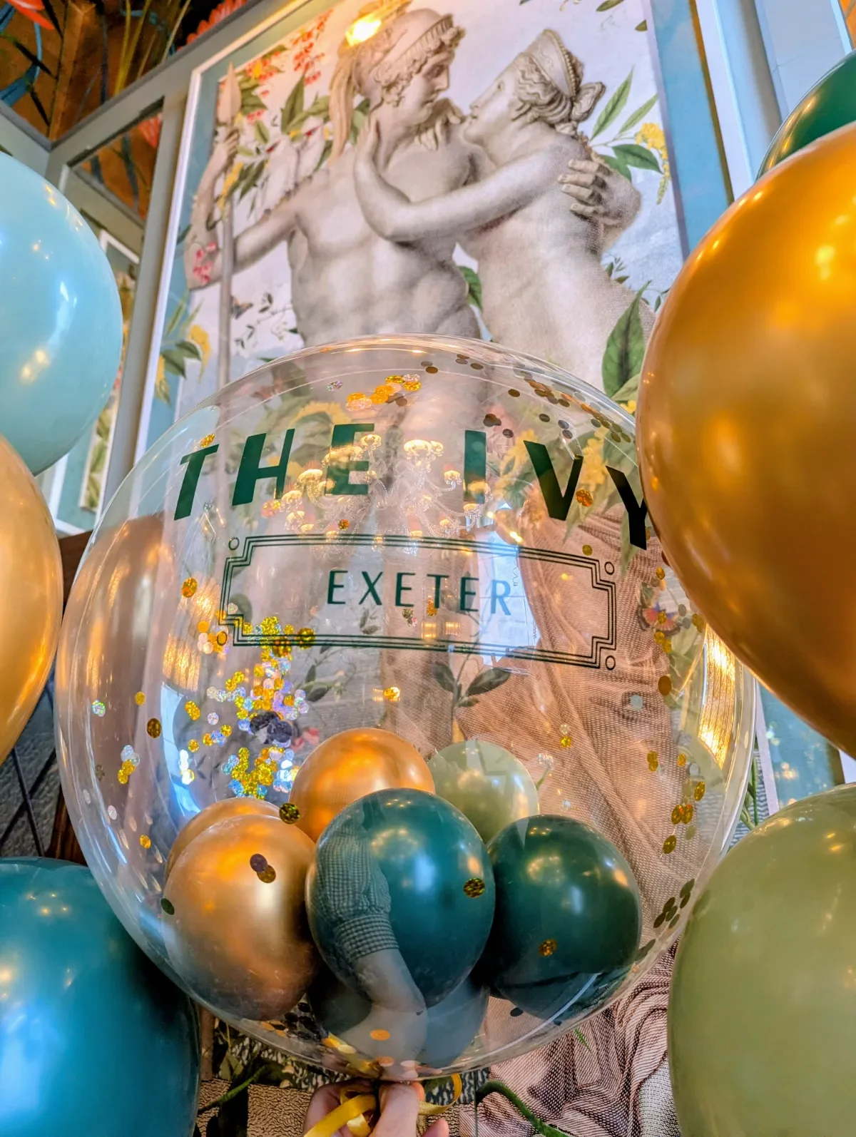 A clear balloon with gold and silver glitter, surrounded by gold, teal, and green balloons, with a background artwork of two classical sculptures of a man and woman embracing, and the text on the balloon reads 'The Ivy Exeter'.