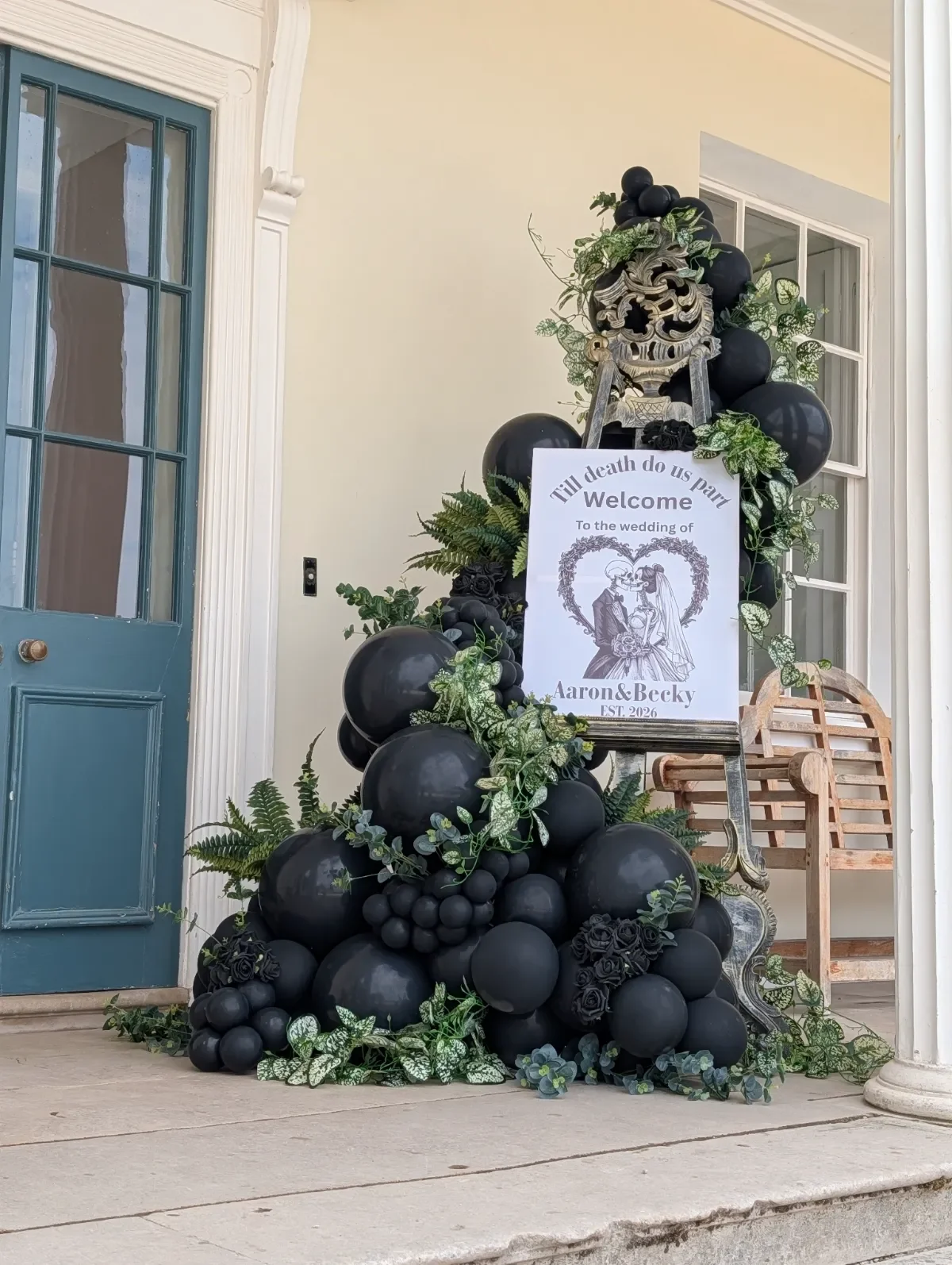 Decorative wedding display with black balloons, greenery, a skeleton-themed sign, and a wooden chair on a porch.