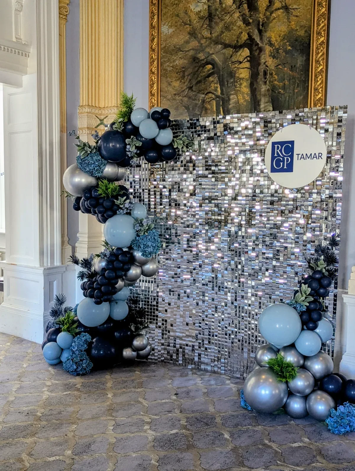 Decorative backdrop with a mosaic mirror panel, balloons in shades of blue, black, silver, and some greenery, with a room featuring ornate architectural details and a large framed painting of a tree in the background.