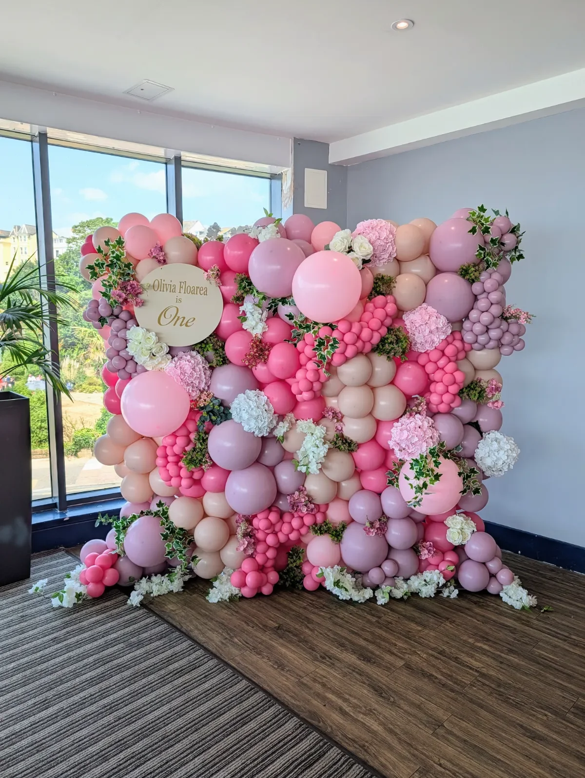 Pink and white balloon display with flowers and a sign that reads "Olivia Floarea is One" for a child's first birthday celebration.