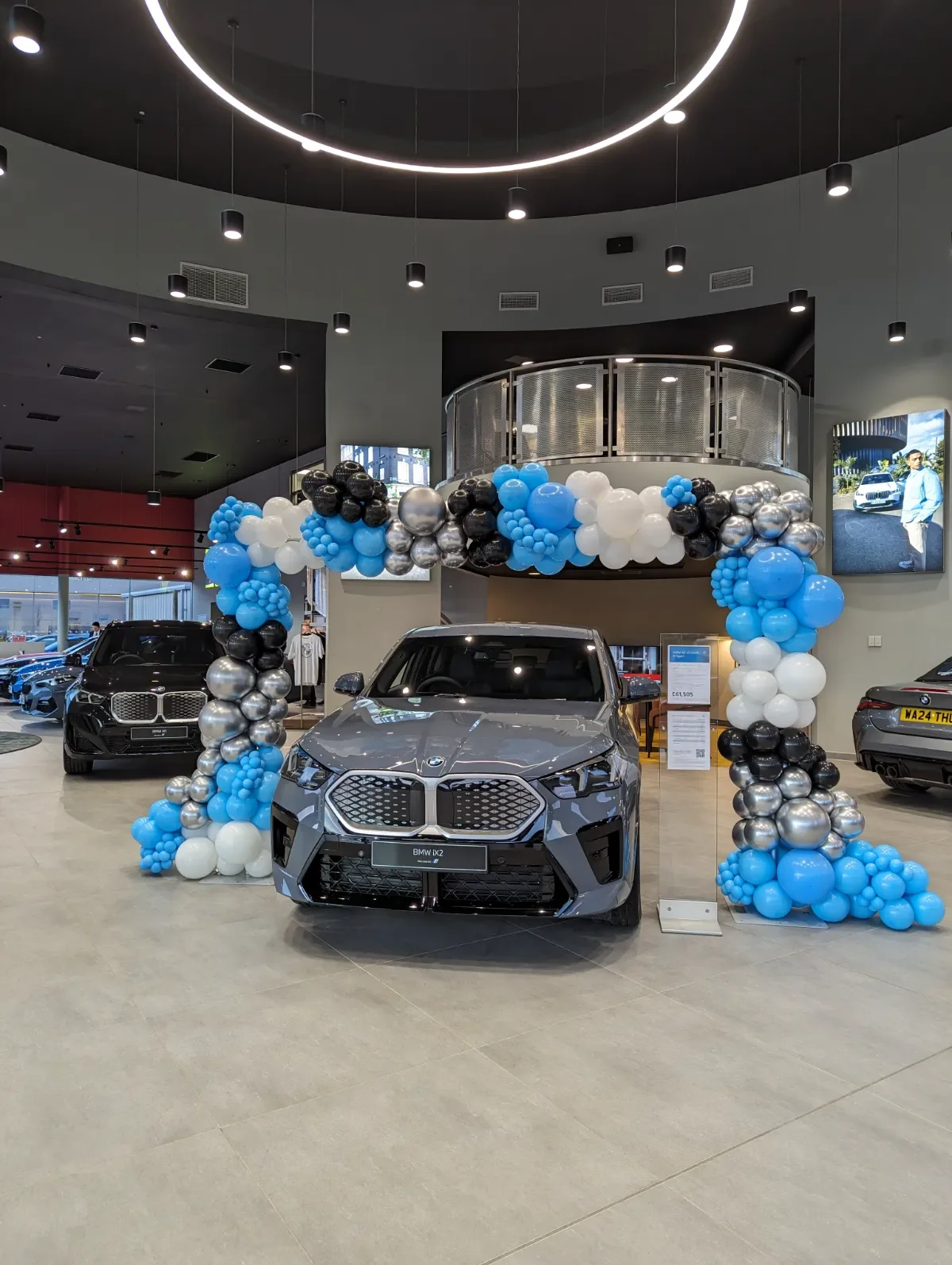 A car dealership showroom with a new gray BMW i2 displayed under a decorative balloon arch in blue, white, black, and silver. Other luxury cars are visible in the background. The showroom has modern lighting and large digital screens on the walls.