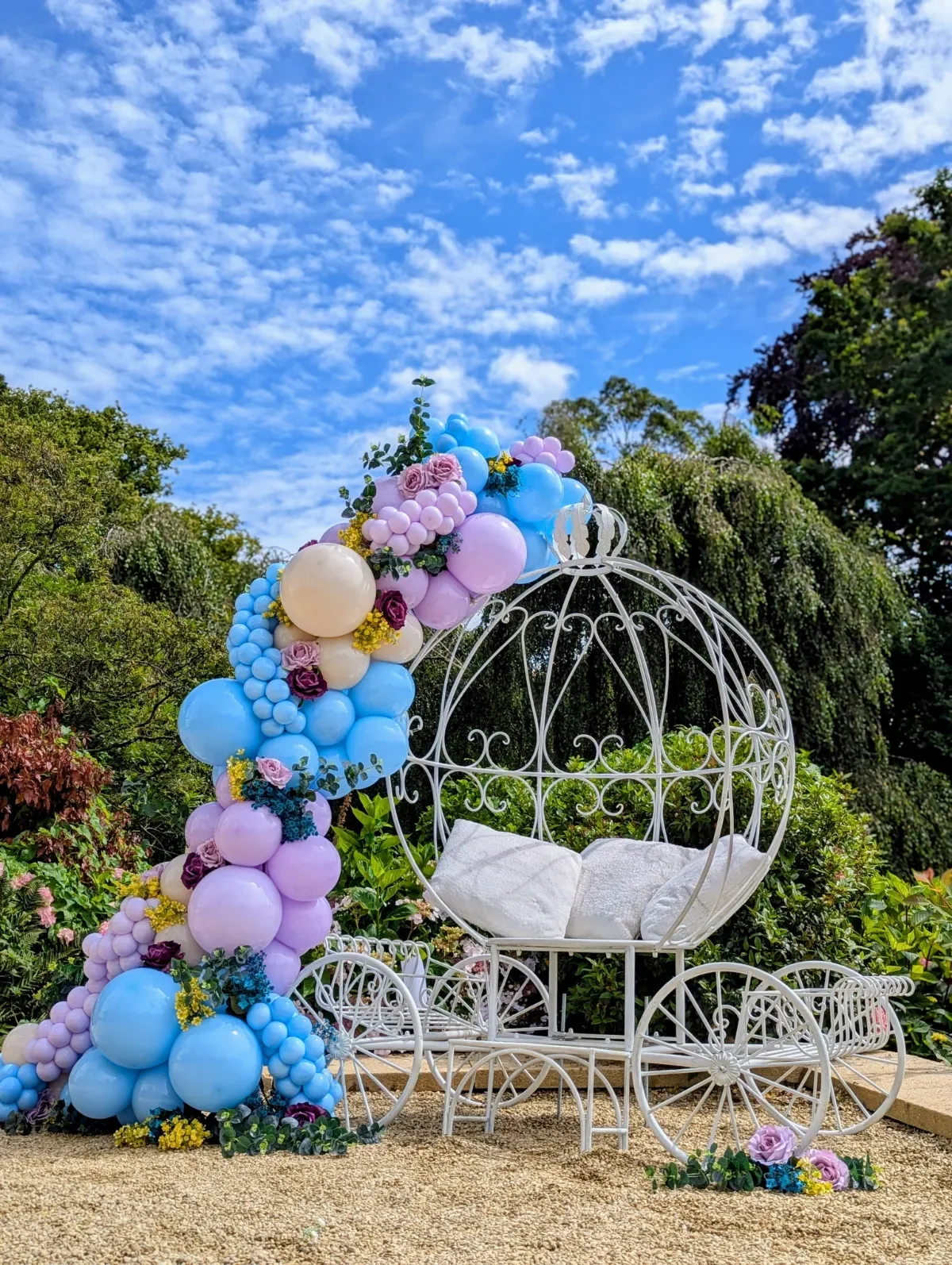 A decorated white, metal carriage with a canopy, surrounded by pastel-colored balloons (blue, pink, and cream), flowers, and greenery, set outdoors on a gravel surface under a blue sky with scattered clouds.
