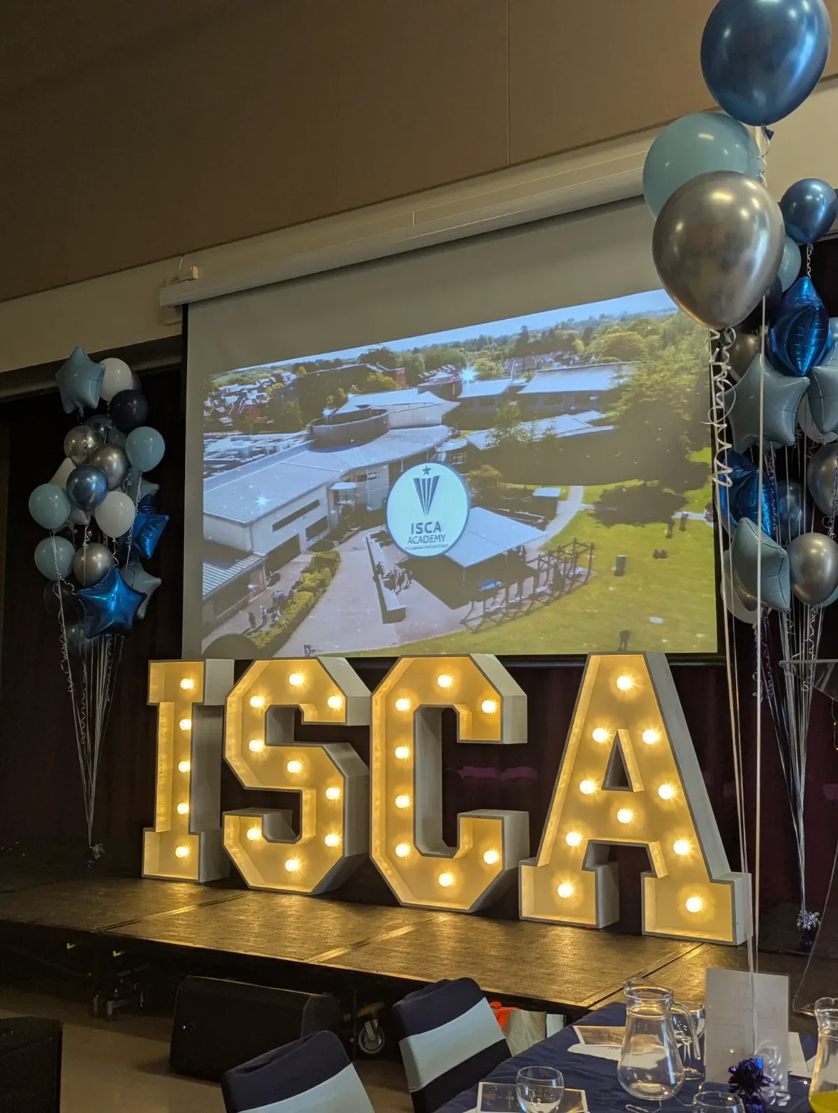 Large illuminated letters spelling 'ISCA' on a stage, decorated with balloons in shades of blue, silver, and white, with a projection screen displaying an aerial view of a school campus with the ISCA Academy logo.