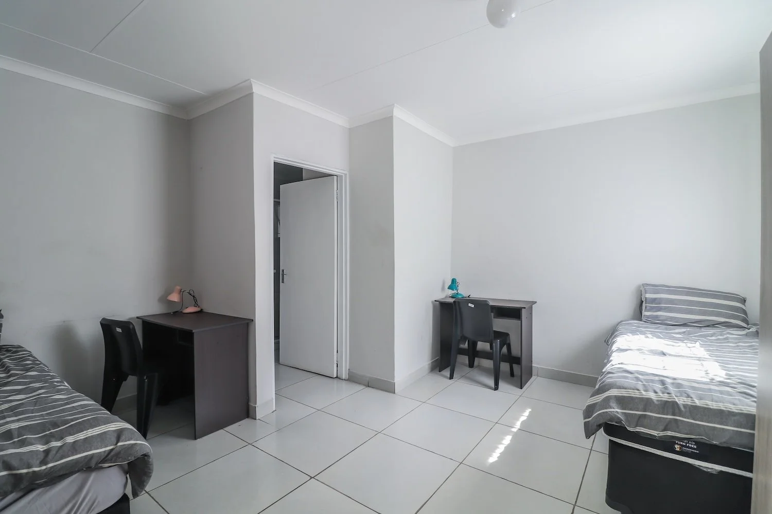 A simple, sparsely furnished bedroom with two single beds featuring gray and white striped bedding, two black desks with chairs, and a plain white tiled floor. One desk has a pink lamp, and the other has a blue desk lamp, with a door slightly open visible in the background.