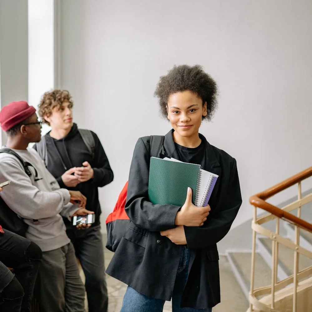 A young woman with afro hair holding notebooks and a backpack, standing on a staircase, smiling at the camera with two other students in the background.