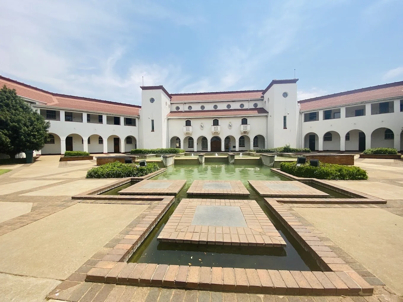 A large white building with a red-tiled roof, arched windows and doors, overlooking a courtyard with a rectangular fountain, green plants, and paved walkways under a partly cloudy sky.