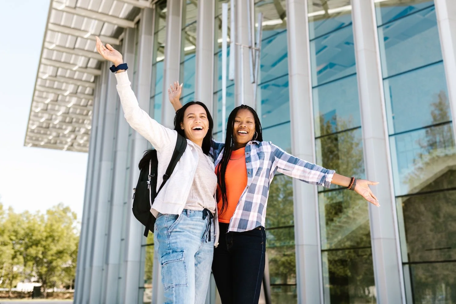 Two young women are smiling and posing happily outside a modern glass building, with arms outstretched. One has a backpack and the other is wearing a plaid shirt and orange top.