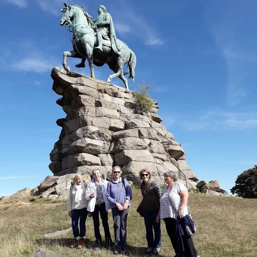 Group of five women standing in front of a large outdoor bronze statue of a man on a horse, mounted on a stone pedestal, under a blue sky.