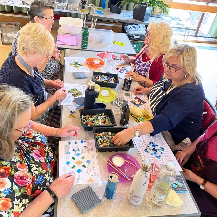 Six women sitting around a table making mosaic art with colored tiles, using instructions and supplies, in a well-lit room.