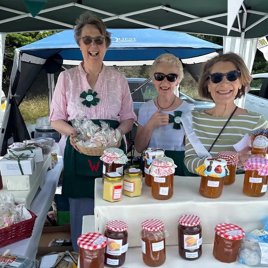 Three women smiling at a stall selling homemade preserves at an outdoor market. They are under a canopy, with jars of jam and honey on display, and some are wearing sunglasses and decorative ribbons.