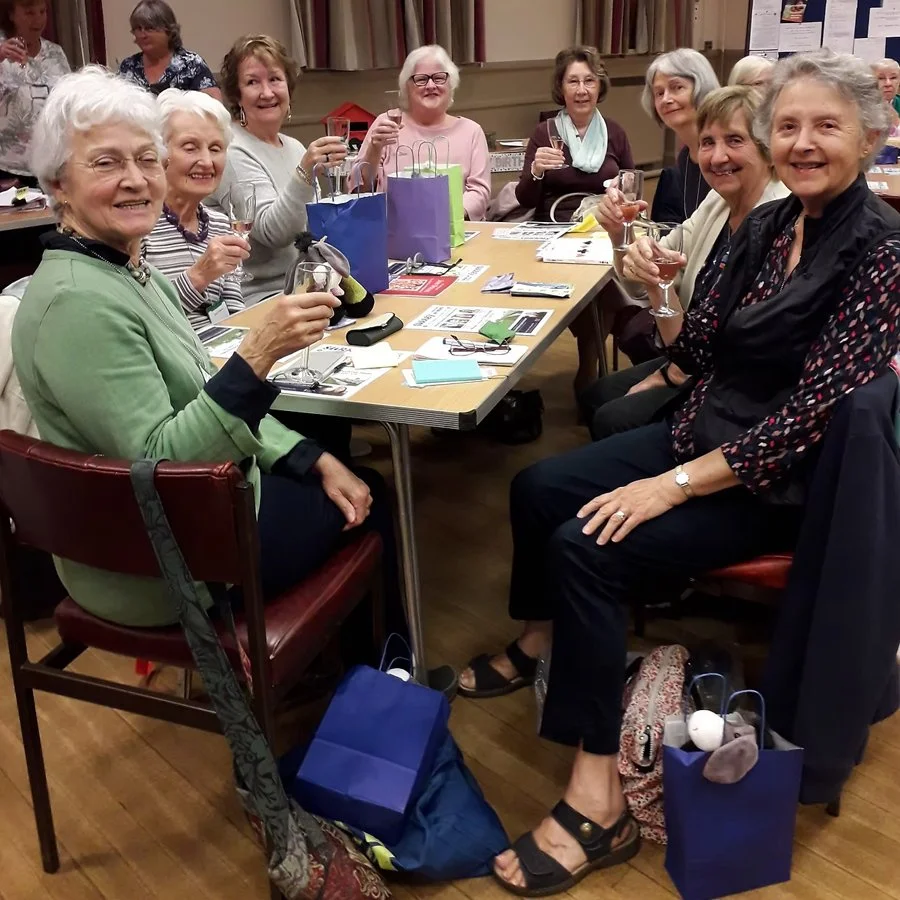 A group of elderly women celebrating at a gathering, sitting around a table with drinks, gift bags, and various items, smiling and toasting glasses.
