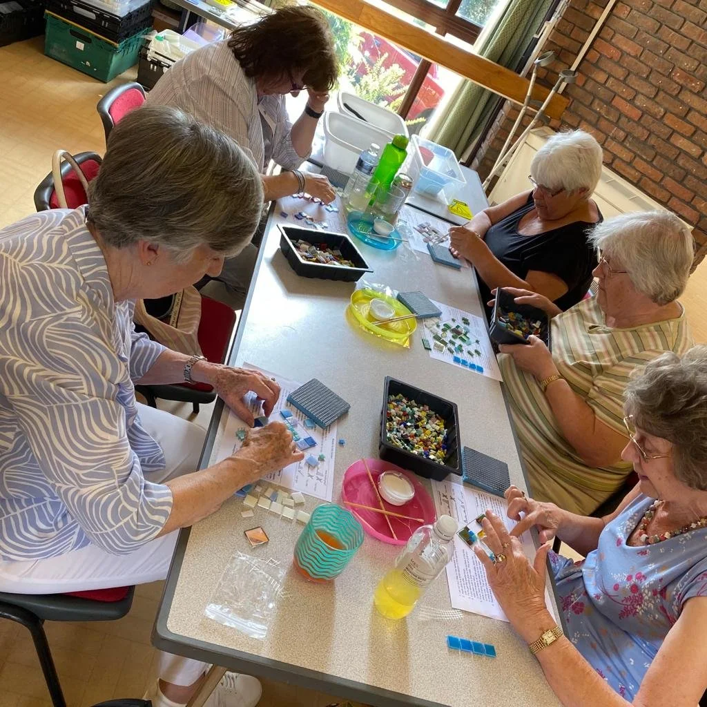 Six women sitting around a table making mosaic art with colored tiles, using instructions and supplies, in a well-lit room.