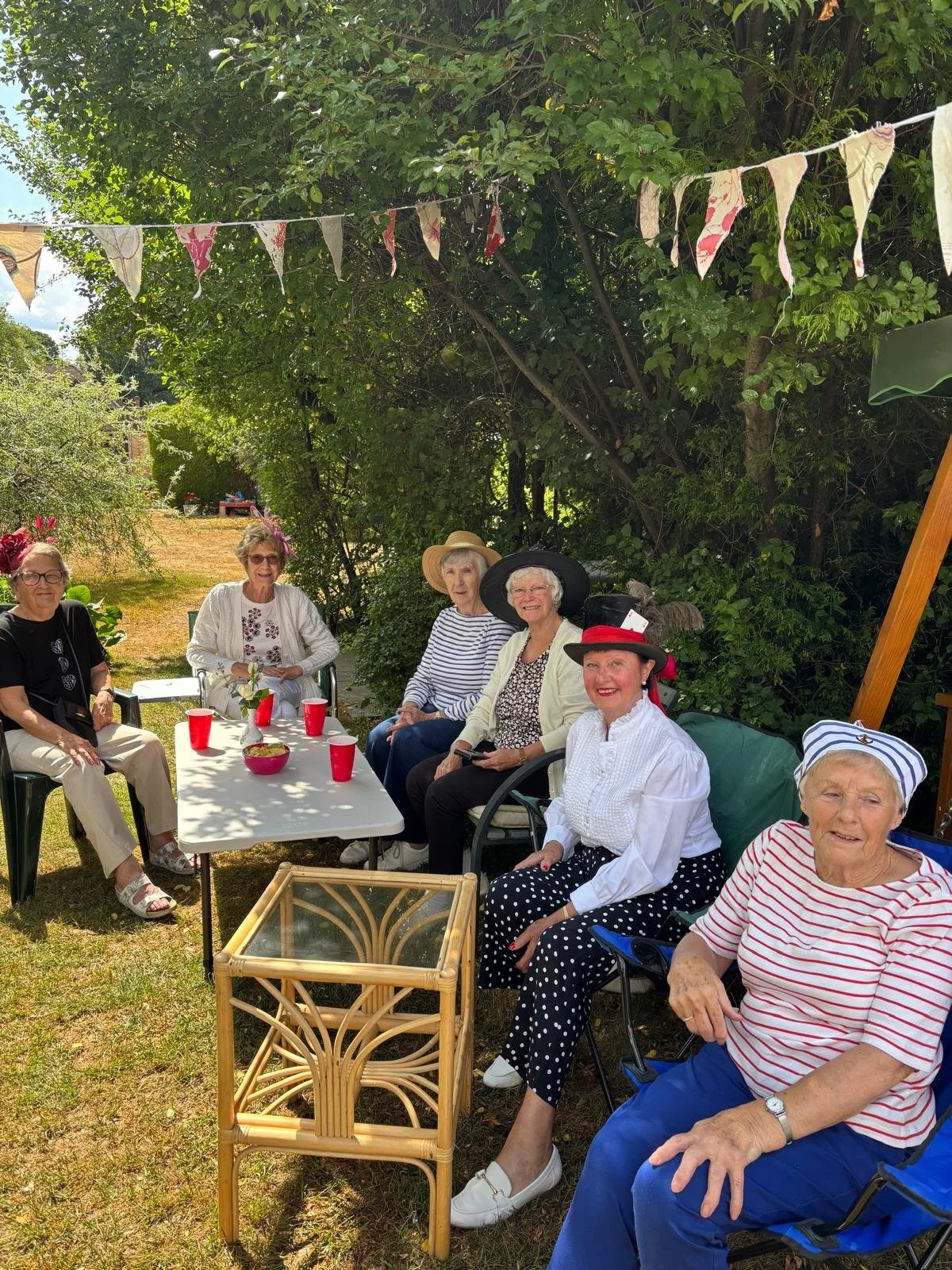 A group of seniors having a gathering outdoors at a garden party. They are sitting around a white table with red cups and a small vase with flowers. The women are wearing hats and casual summer clothes, smiling in a shaded area with greenery and bunting decorations overhead.