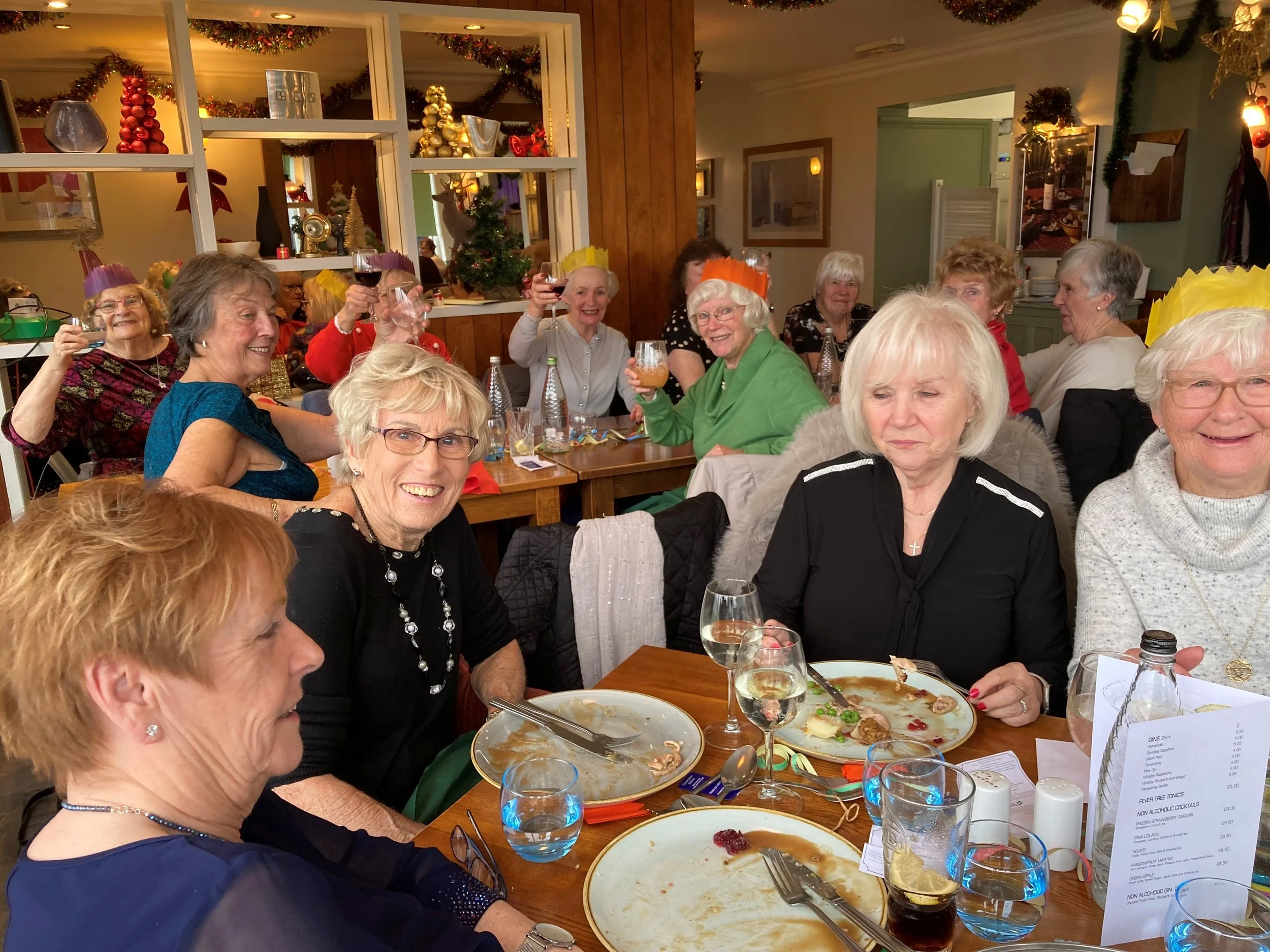 A group of elderly women celebrating Christmas, wearing colorful paper crowns, raising glasses of wine, and smiling at a decorated indoor restaurant with Christmas ornaments and a small Christmas tree in the background.