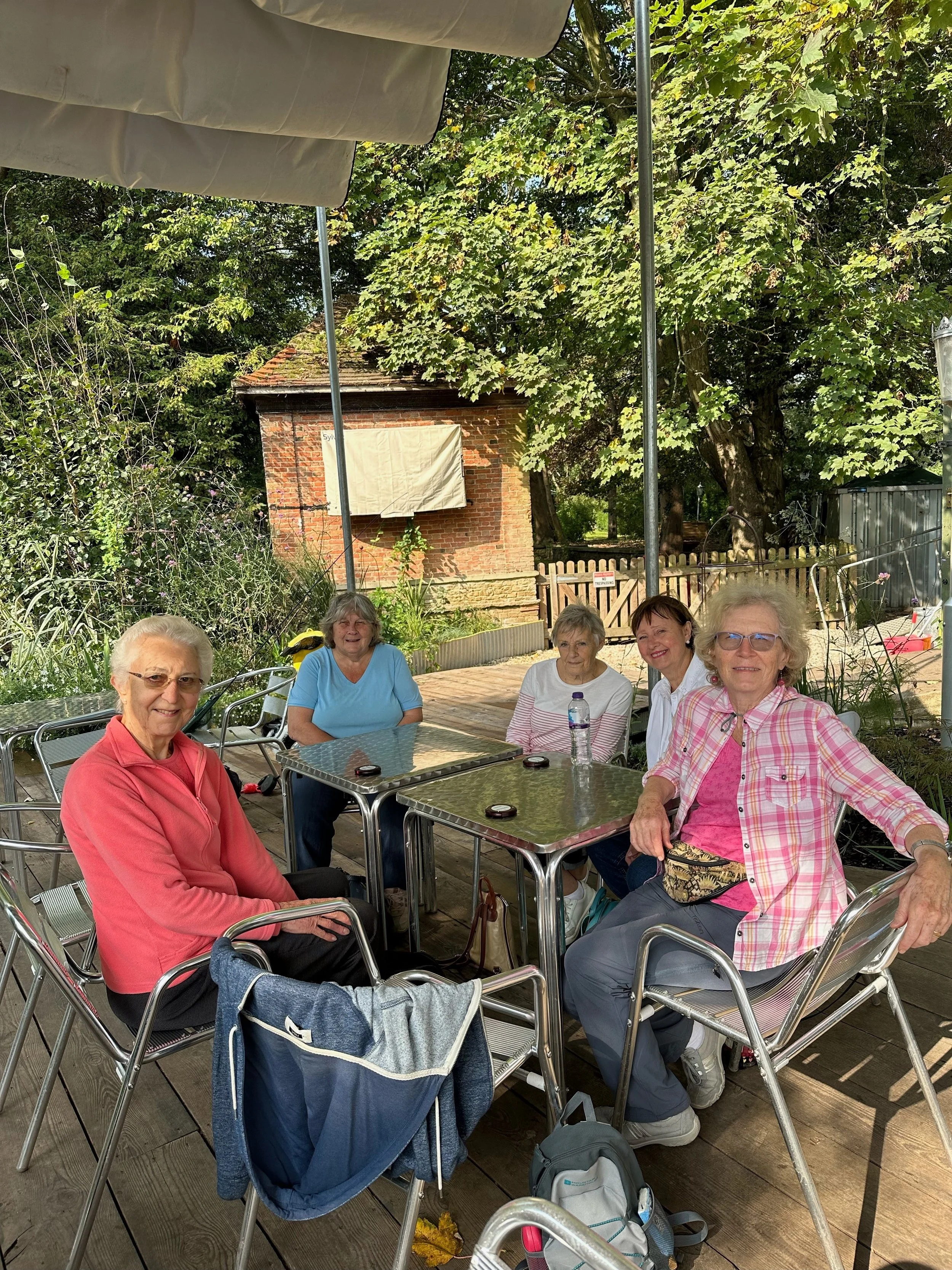 A group of five elderly women sitting at outdoor tables under large beige umbrellas, enjoying a sunny day among trees and plants in a garden setting.