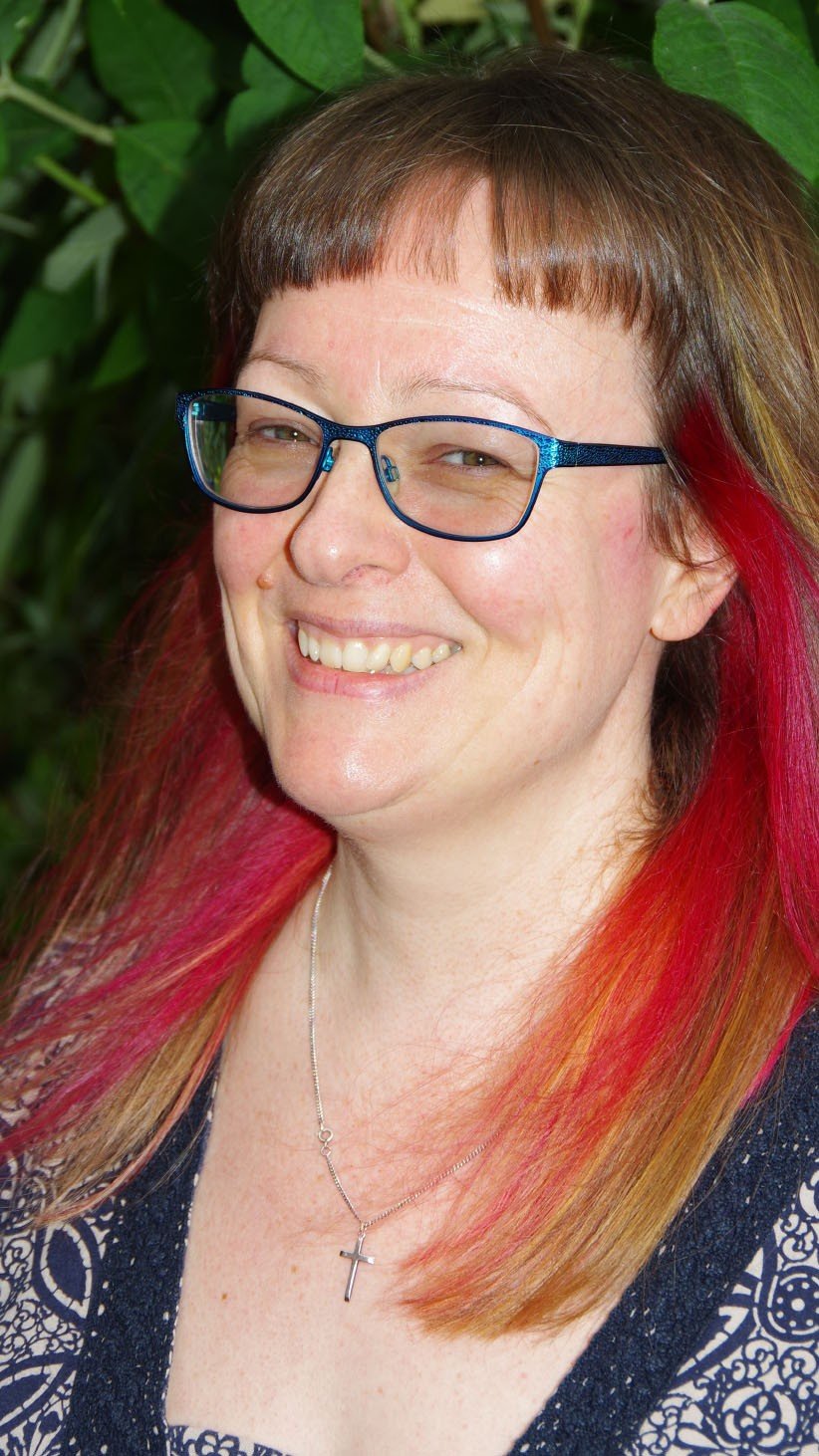 A woman smiling outdoors, wearing glasses, with reddish hair and a necklace with a cross, with green leaves in the background.