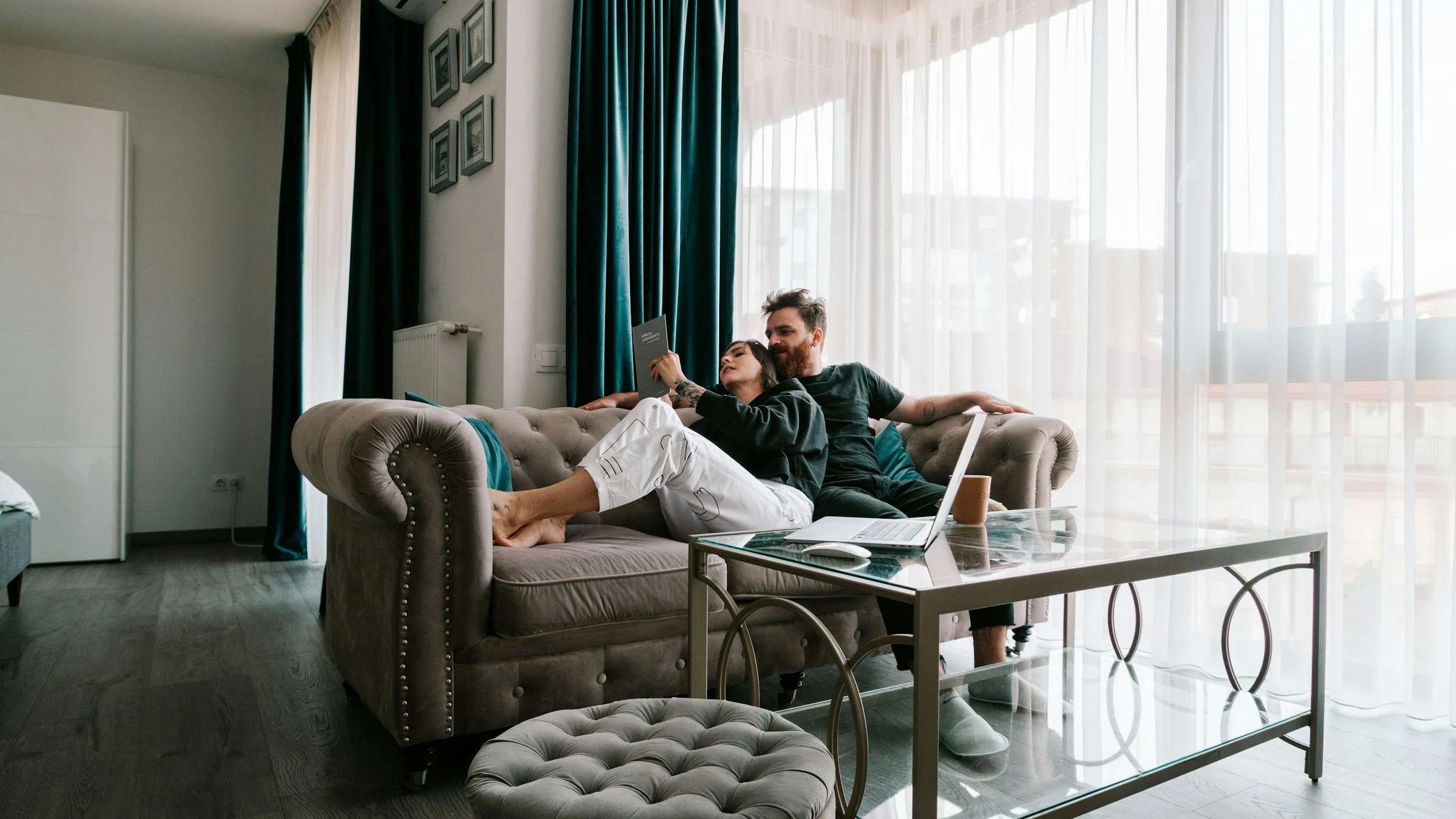 A man and a woman sitting together on a sofa in a living room, with the woman reading a book and the man sitting close to her, surrounded by a laptop, coffee mug, and a glass table, near large windows with curtains.