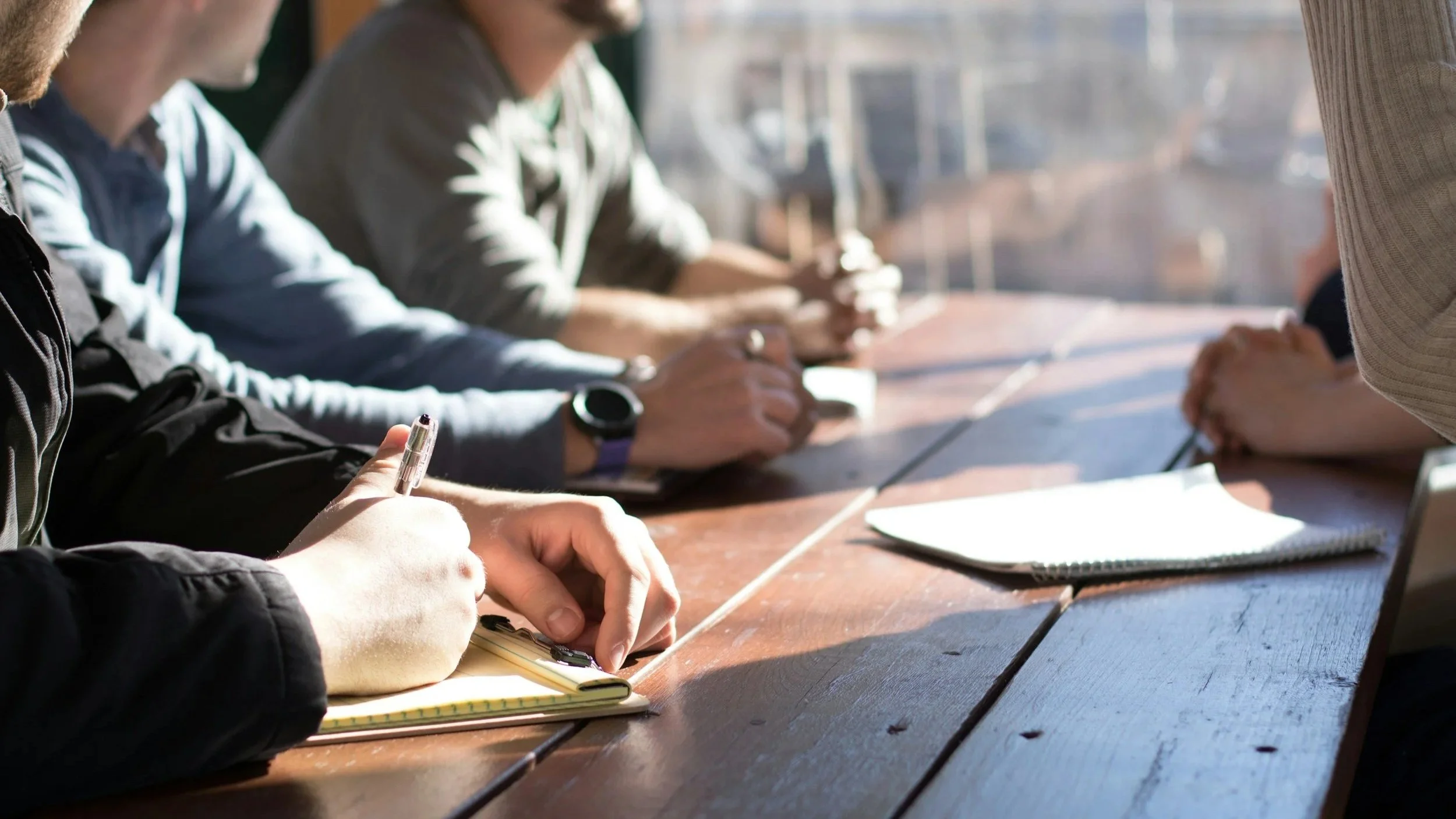 People sitting at a wooden table, writing, drawing, and using notebooks and papers.