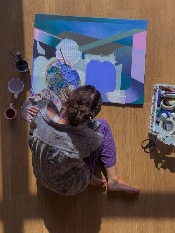 An artist sitting on the wooden floor, painting an abstract landscape on a canvas. The artist's supplies, including paintbrushes and jars of paint, are nearby.