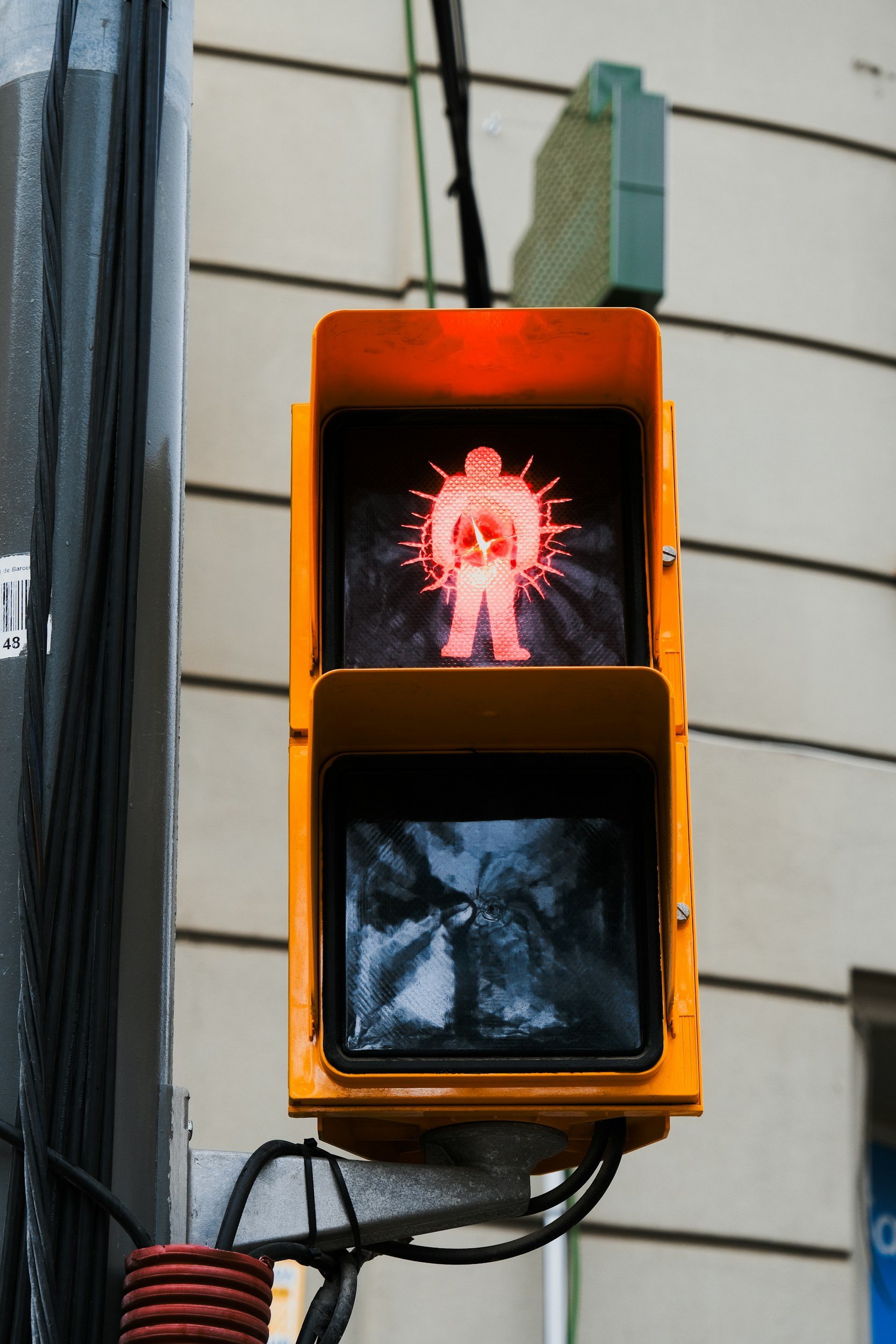 A traffic signal with a red pedestrian figure illuminated, indicating pedestrians should stop. The signal is mounted on a pole with black cables and a red coiled wire at the bottom. The background features a beige building with horizontal siding.