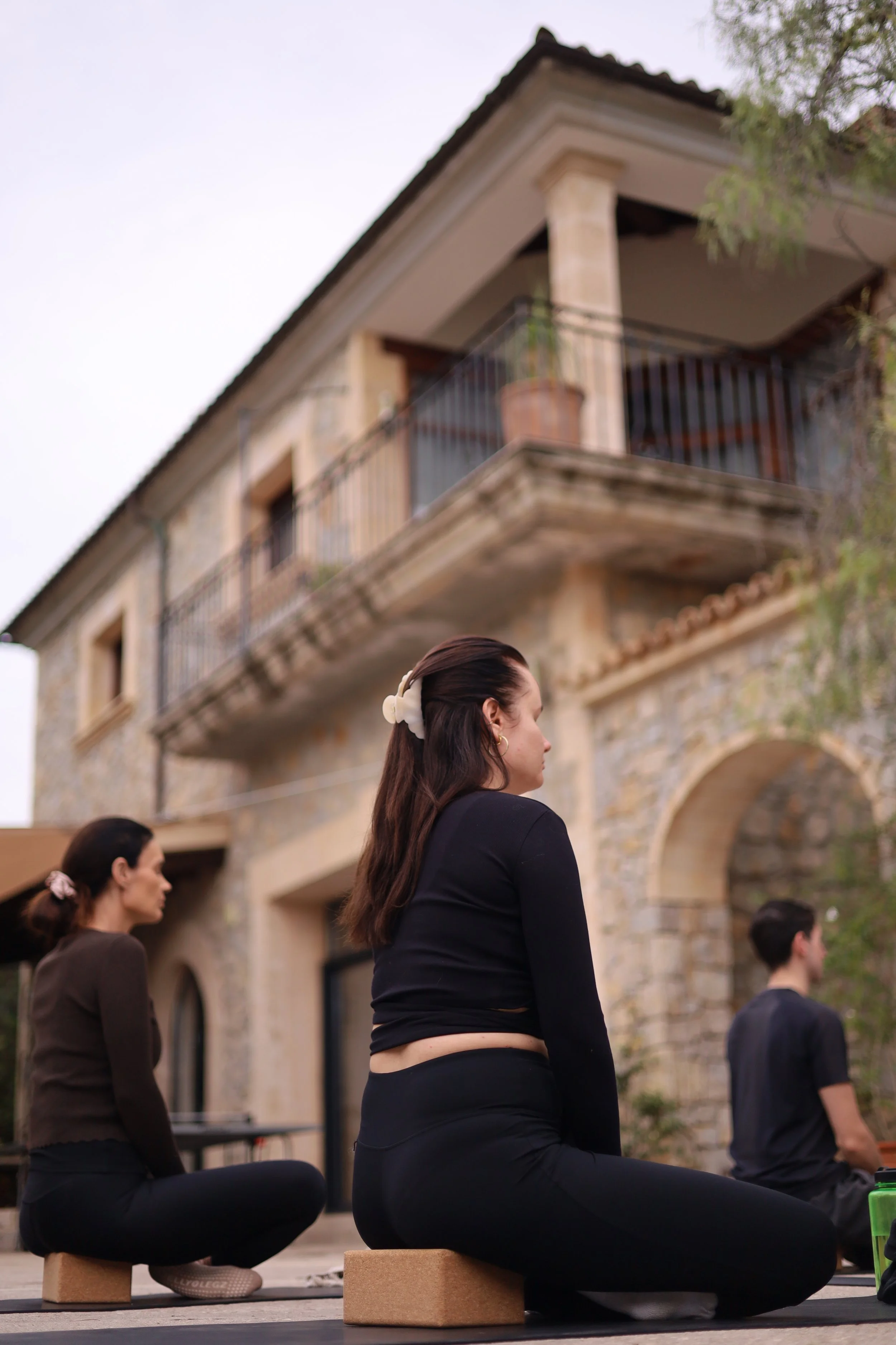 People practicing yoga outdoors on a stone patio, sitting on yoga blocks with a stone building in the background.