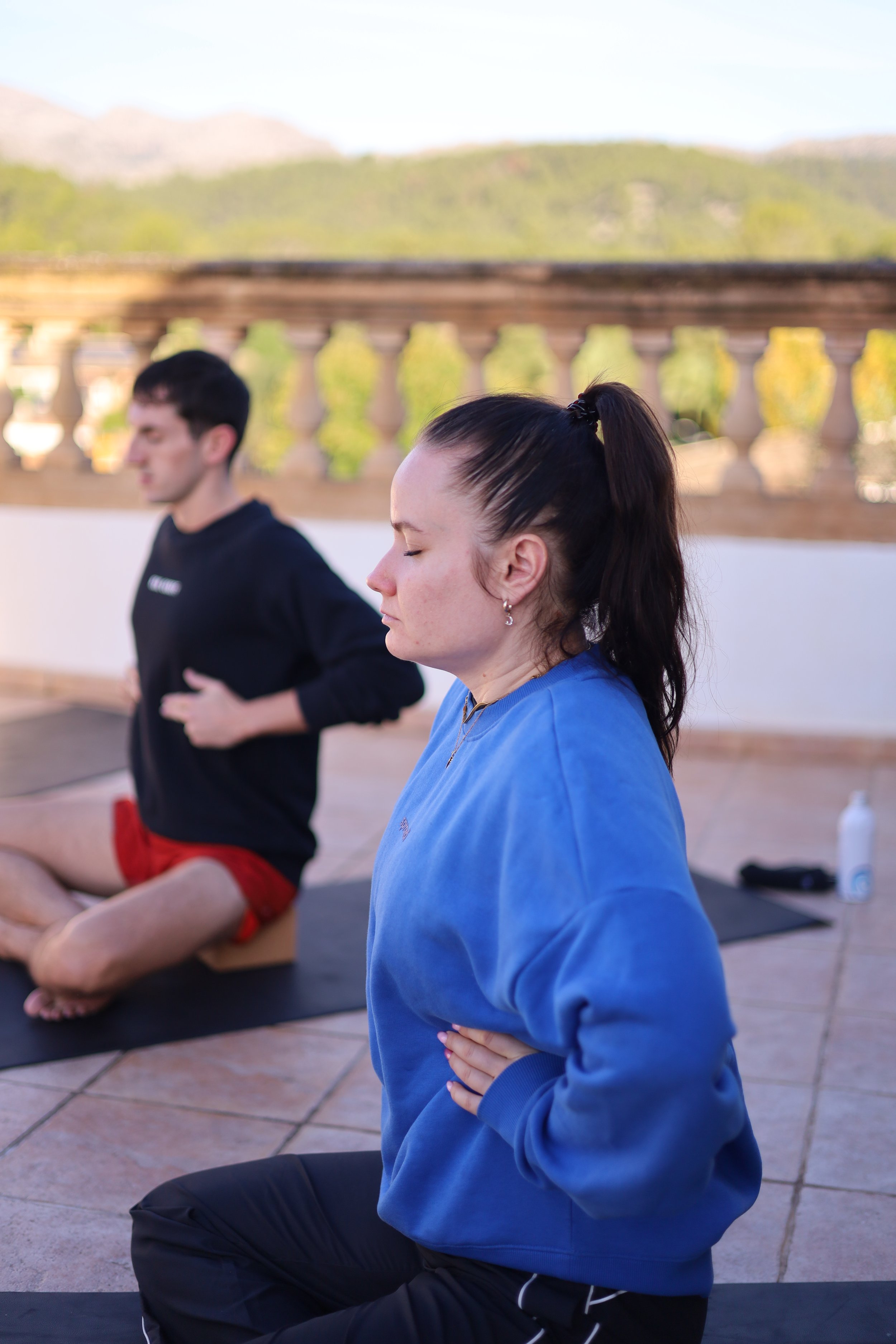 A woman with dark hair in a ponytail practicing meditation outdoors with eyes closed, eyes closed, wearing a blue sweatshirt and black pants, while a man with short dark hair sits cross-legged in the background, wearing a black shirt and red shorts, on a patio with a mountain and greenery in the background.