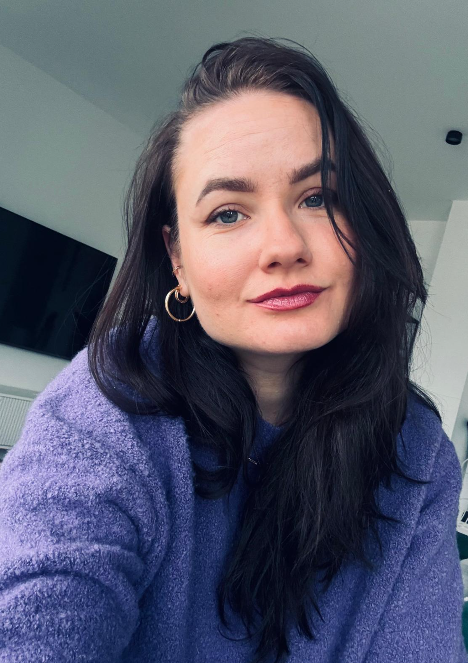 A woman with long dark hair, wearing a purple sweater and gold hoop earrings, taking a selfie in a modern indoor setting.