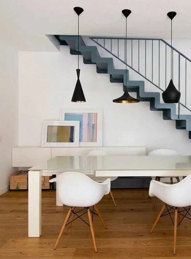 Sala da pranzo moderna con tavolo bianco e sedie bianche con gambe in legno, sopra un tavolo a soffitto ci sono tre lampade nere, e vi è una scala a zig zag color blu con ringhiera nera sullo sfondo, decorazioni minimaliste con quadri appoggiati su una panca bianca.