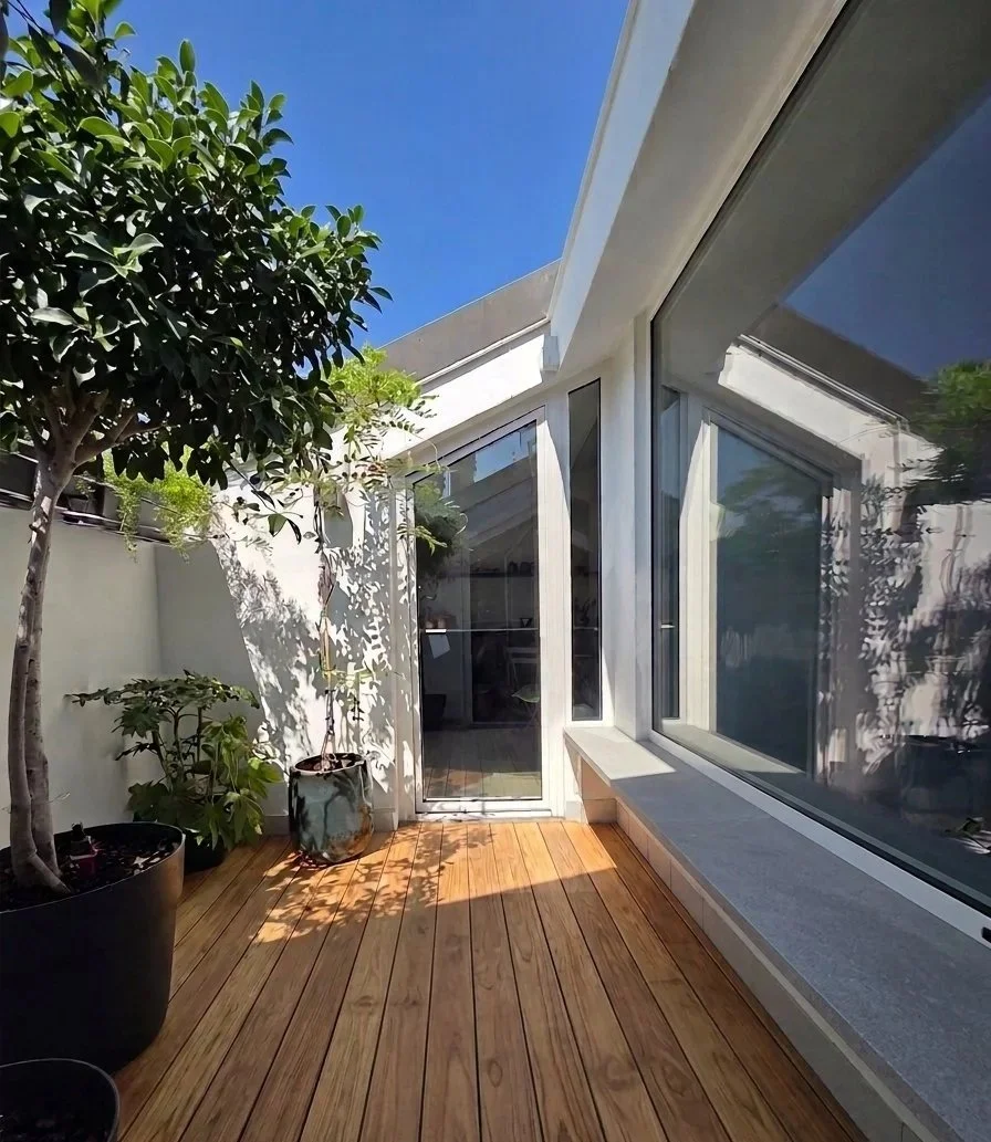 Terrazza con pavimento di legno, piante in vasi grandi e piccole, muro bianco e grandi finestre con riflesso del giardino, sotto un cielo azzurro.
