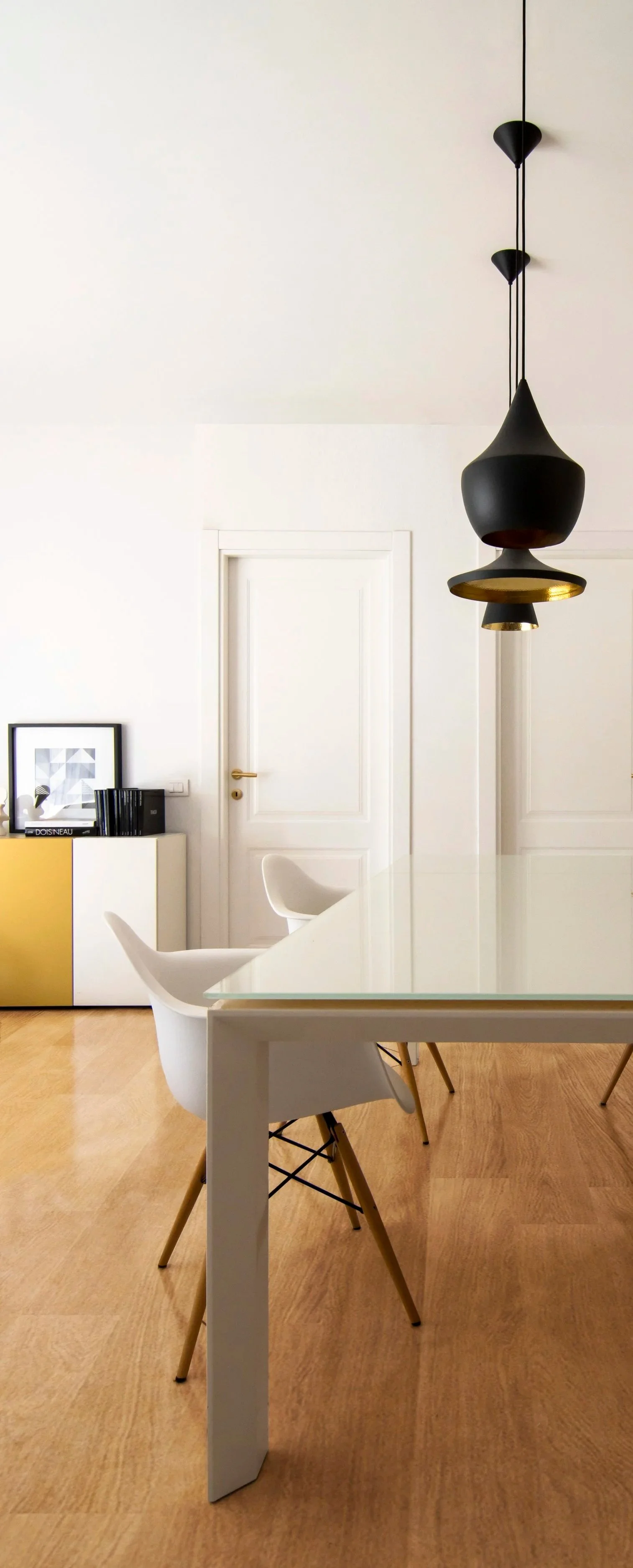 Interno di una stanza moderna con tavolo bianco, due sedie bianche con gambe in legno, lampadario tom dixon nero sospeso, porta bianca e pareti chiare, pavimento in legno.