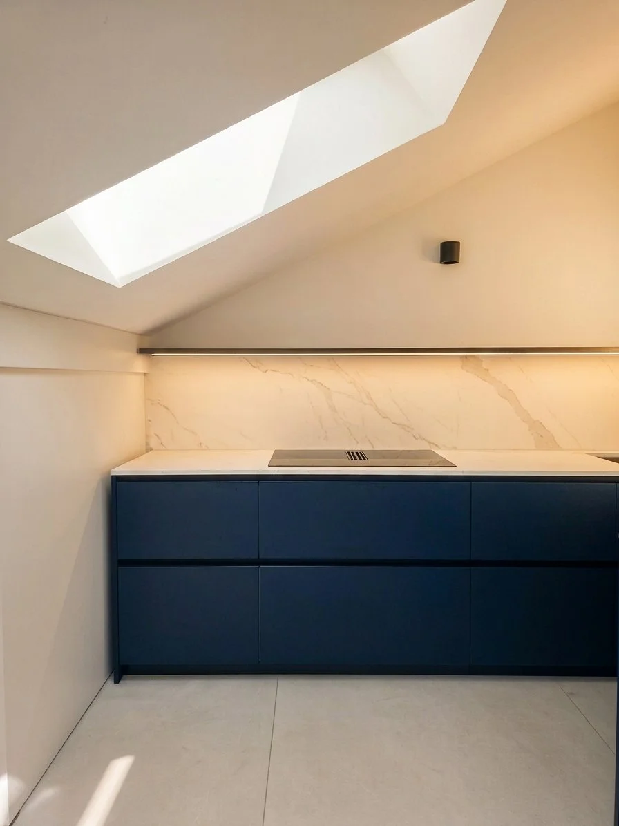 Cucina moderna sotto un soffitto a soffitto inclinato con finestra skylight, armadietti blu, piano bianco e parete in marmo