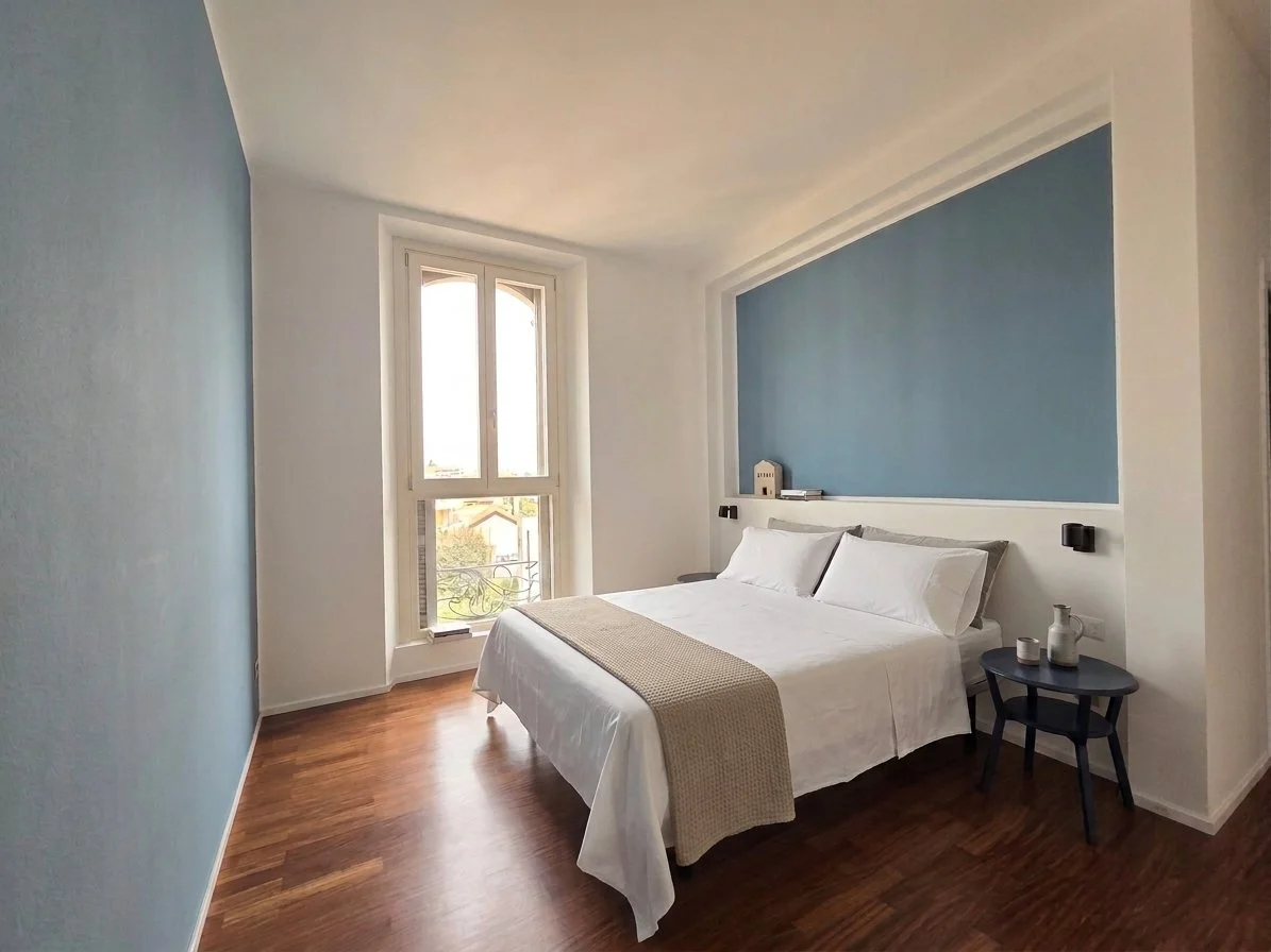 Camera da letto con letto matrimoniale, cuscini bianchi, pareti bianche e una parete blu sopra il letto, finestra con vista esterna, pavimento in legno, tavolino nero con oggetti decorativi.