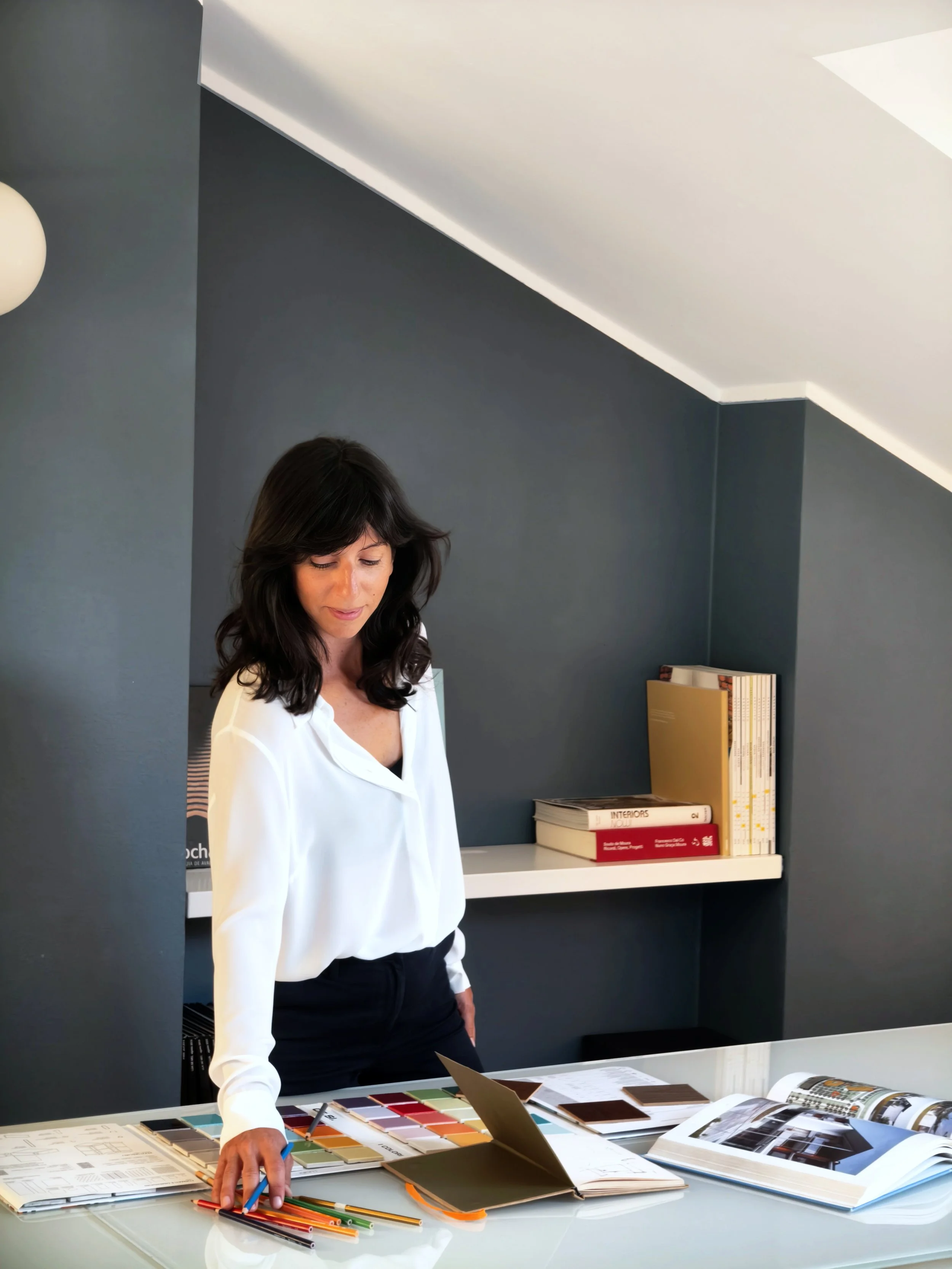 Una donna con capelli castani e vestito bianco si trova davanti a un tavolo con campioni di colori, libri di design e schizzi, in uno studio moderno con pareti grigie e scaffali con libri.