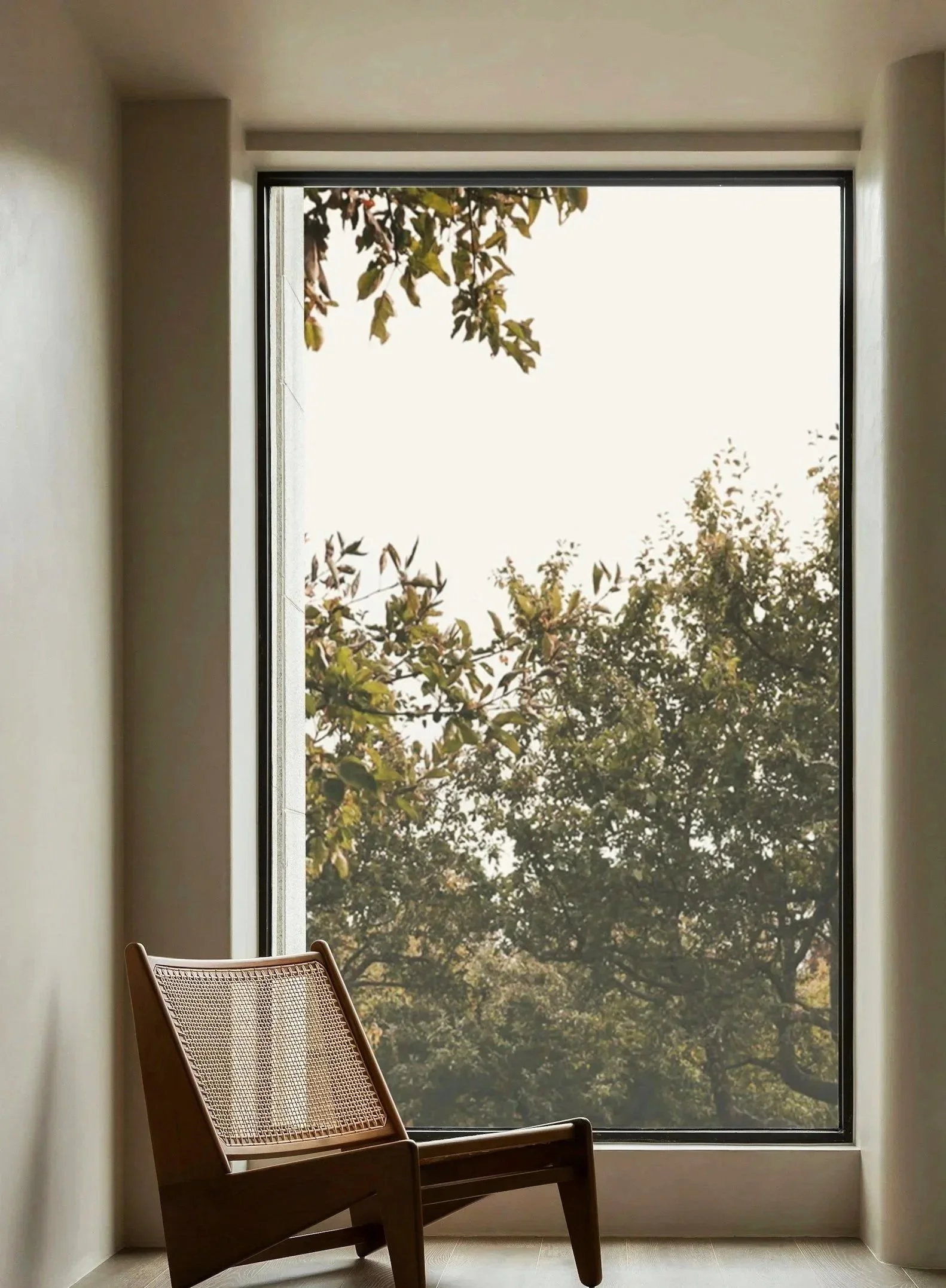 Una sedia di legno con seduta in cesti di paglia si trova davanti a una grande finestra con vista su alberi con foglie verdi e marroni, all'interno di una stanza luminosa con pareti bianche.