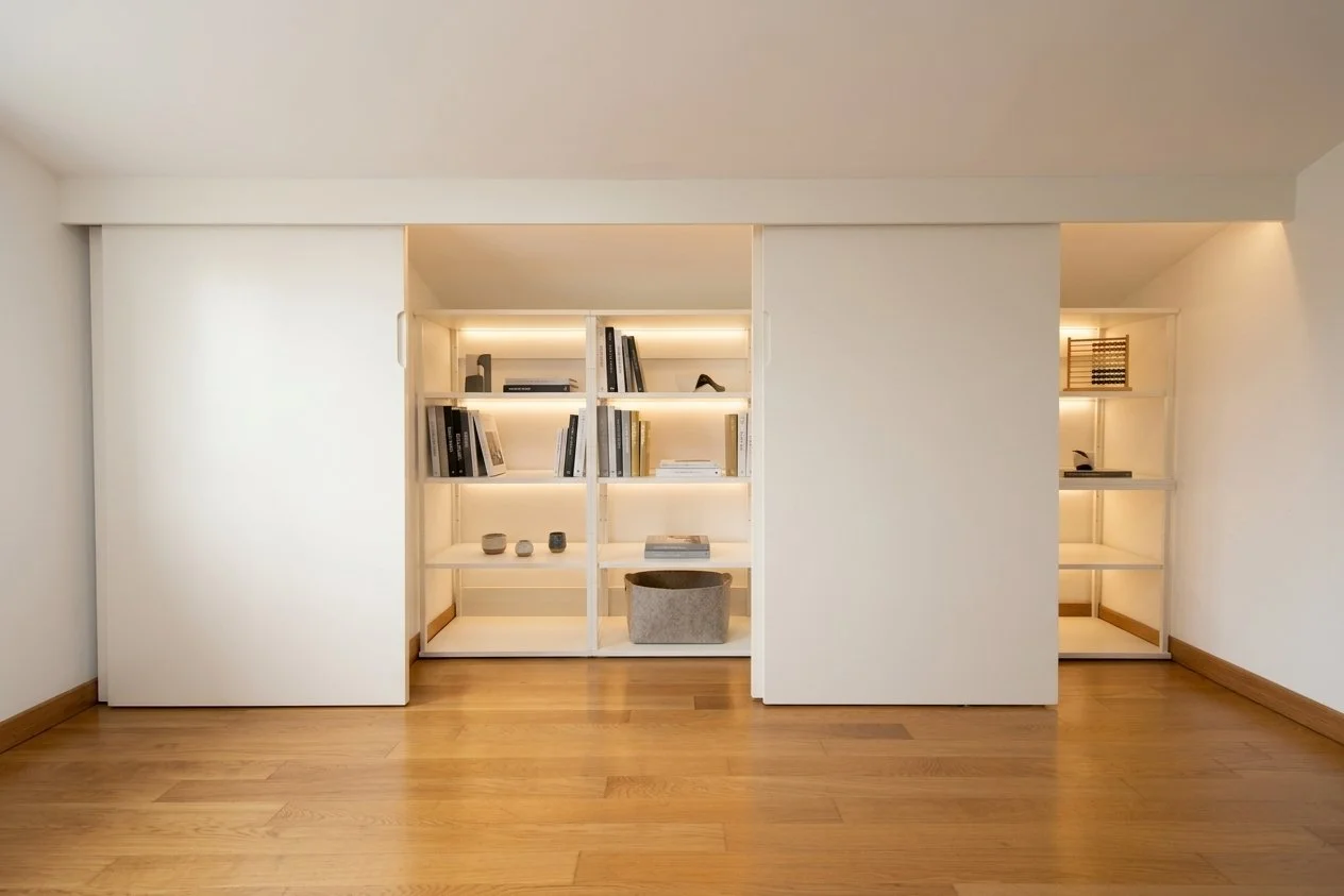 Libreria moderna con scaffali bianchi, alcuni ripiani illuminati, alcuni libri, oggetti decorativi e una scatola di tessuto, con pavimento in legno e pareti bianche.